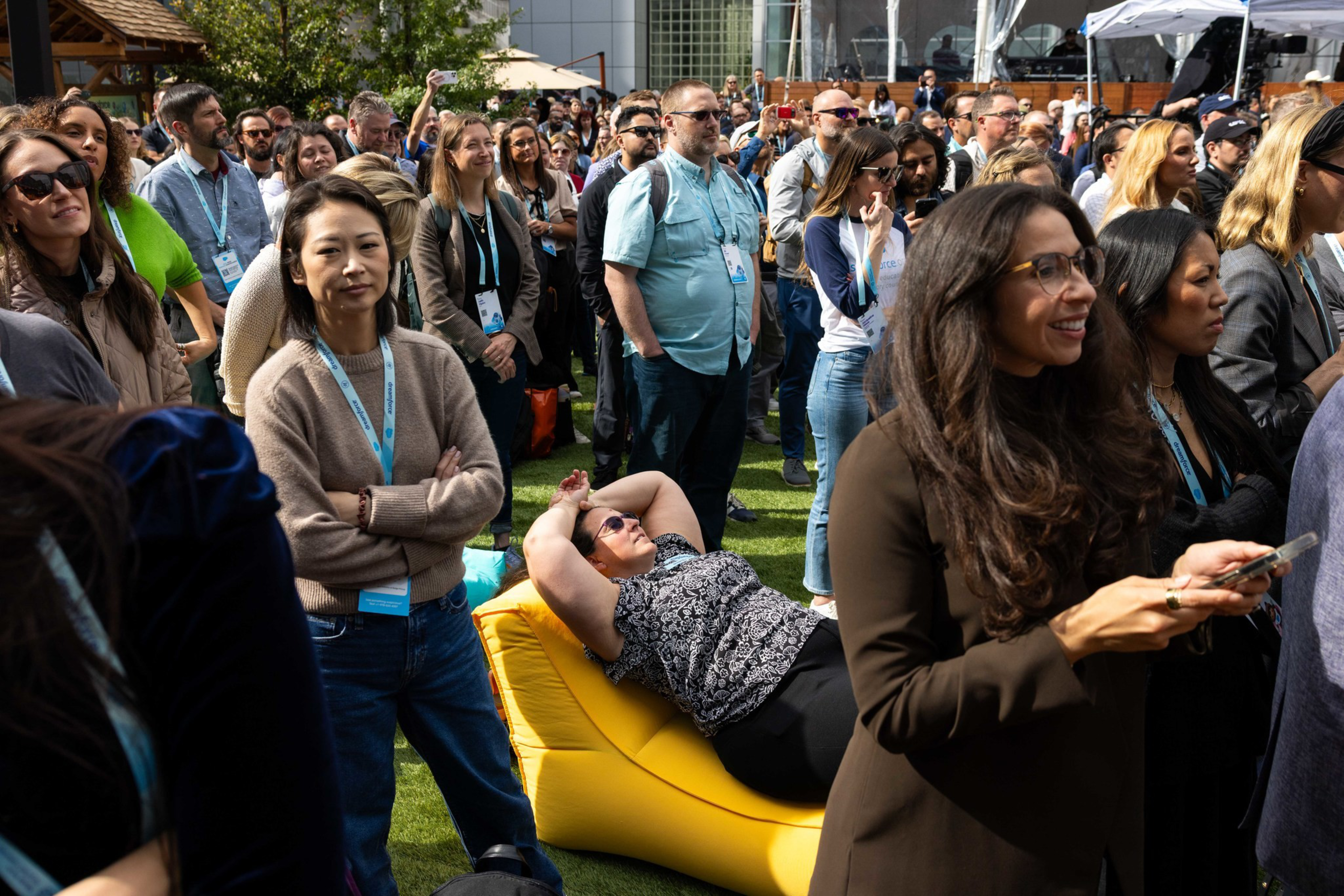 A large group of people stand closely together outdoors on grass while one woman reclines on a yellow inflatable lounger, wearing sunglasses.
