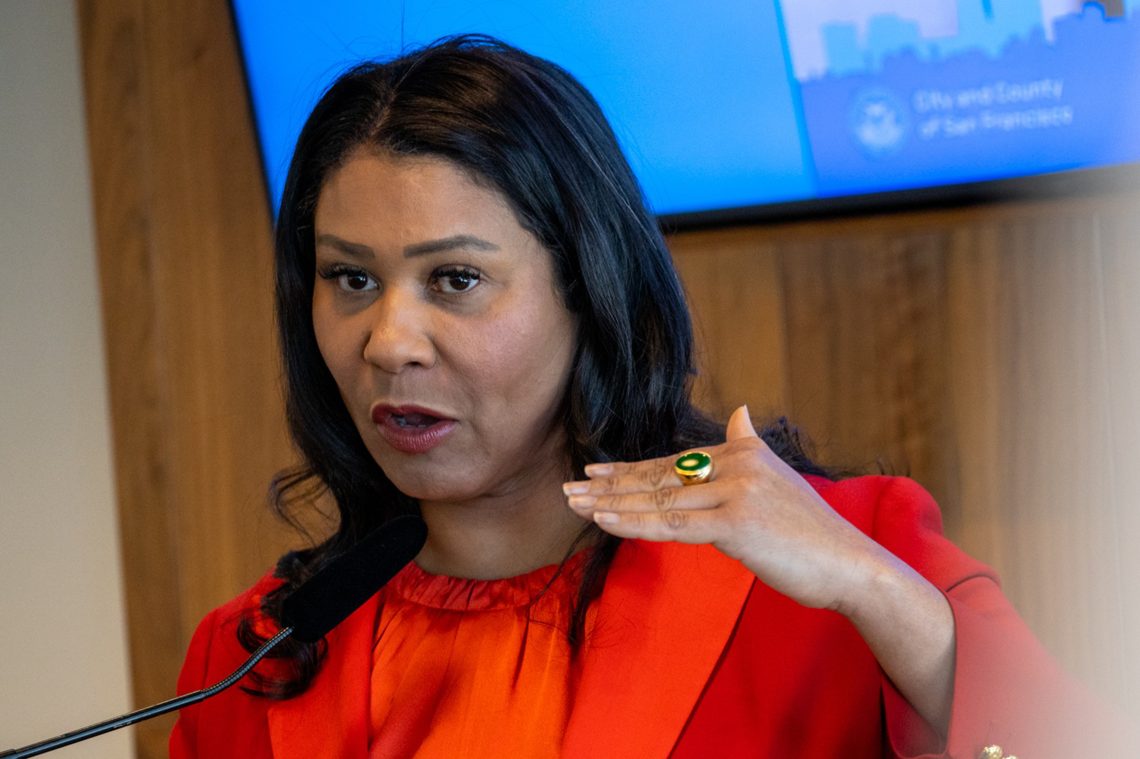 A woman with long dark hair wearing a red outfit speaks while raising one hand near her face, wearing a green ring on her finger.