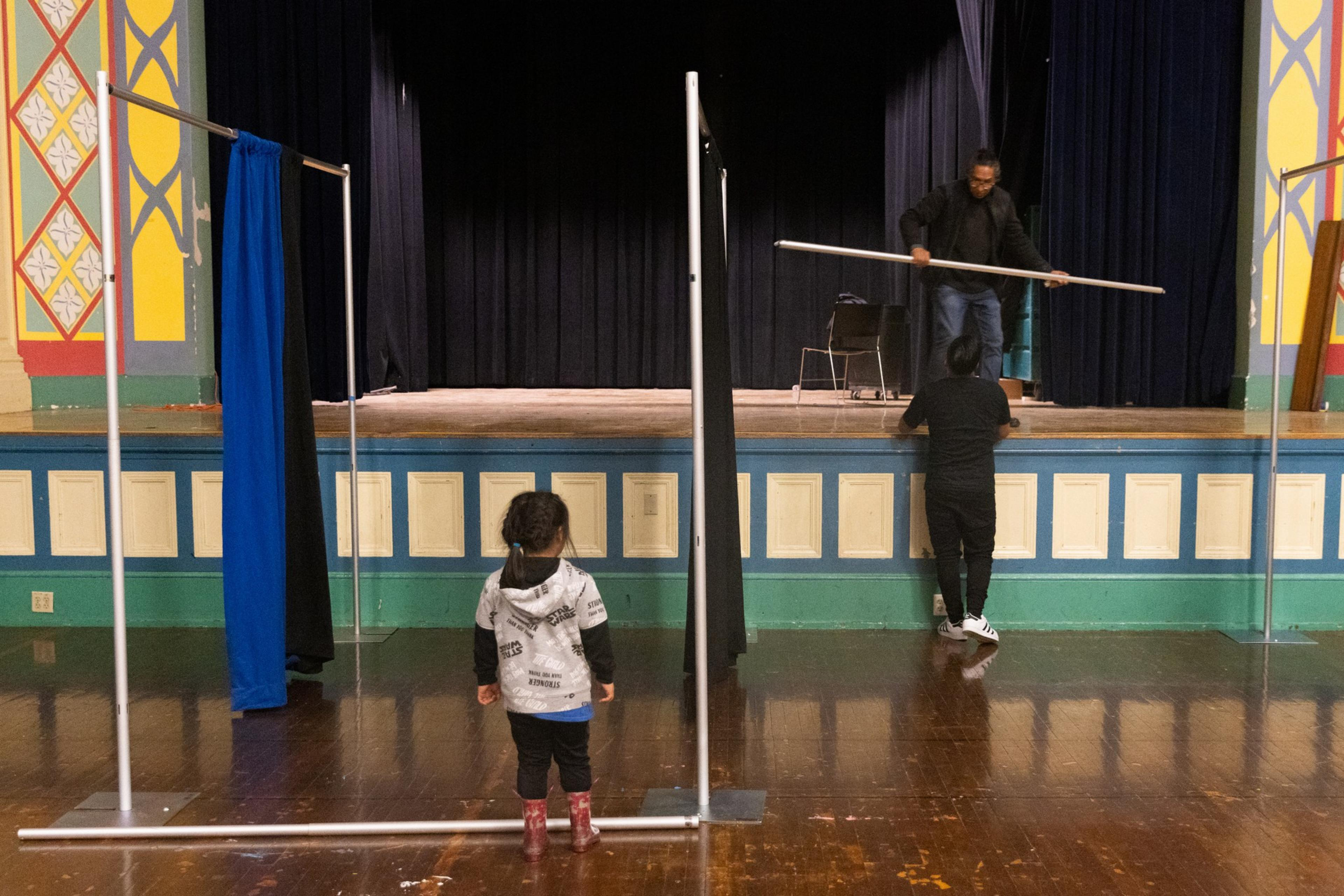 A child watches as two adults assemble privacy dividers in a school gym.