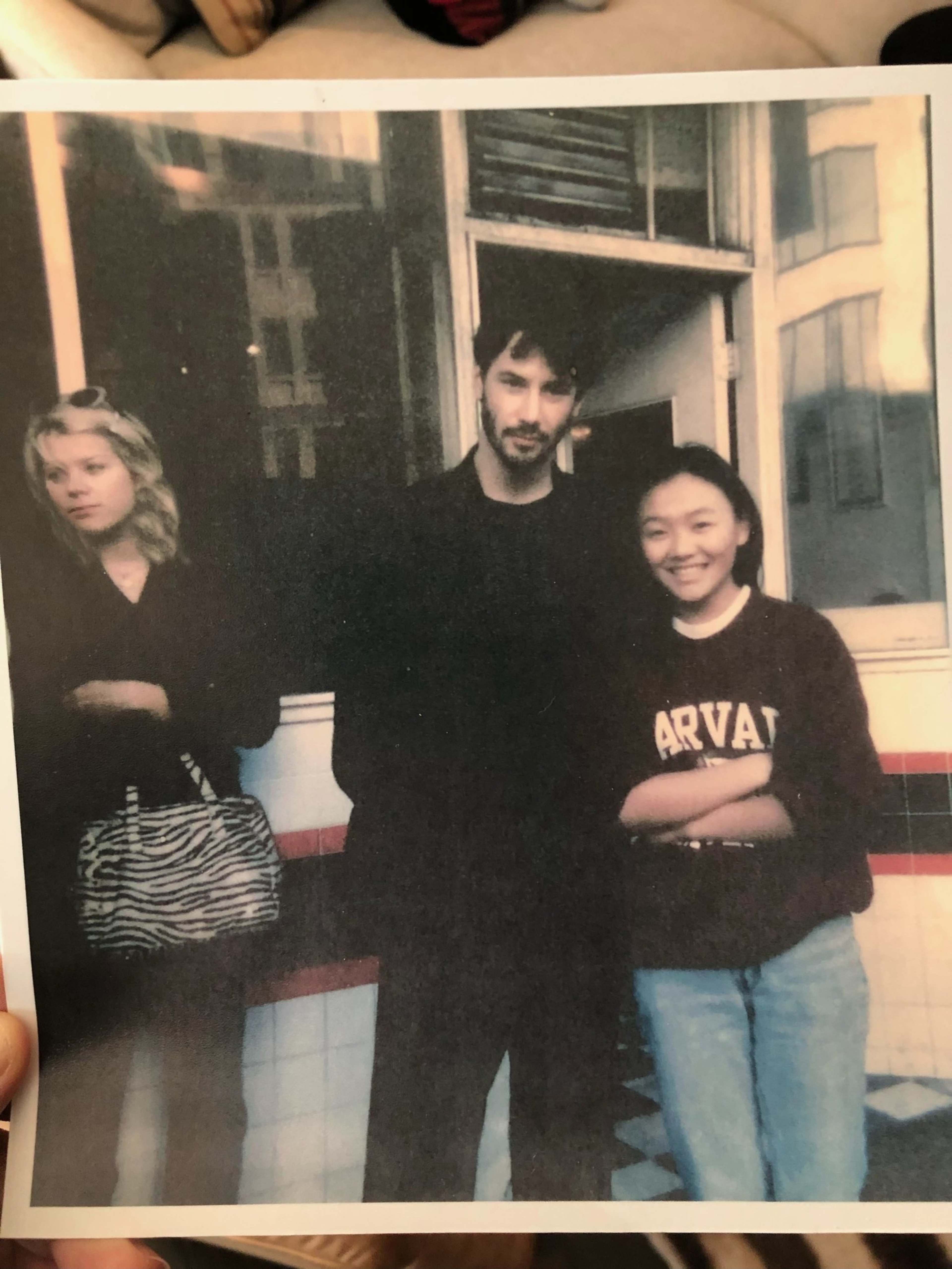 Three people stand outside a building with tiled walls; a man in black is centered between two women, one wearing a Harvard sweatshirt and the other holding a zebra-striped bag.