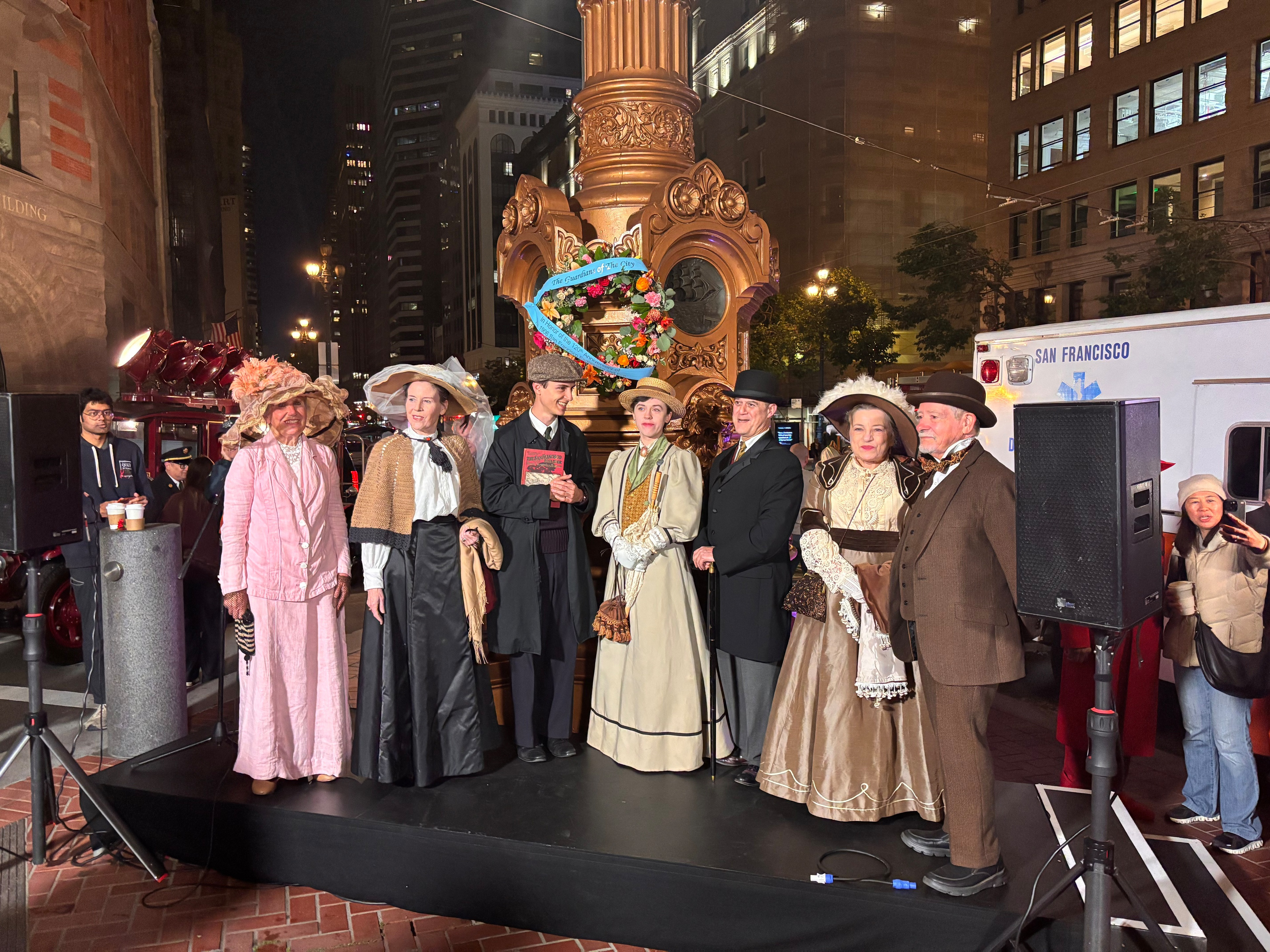 Seven people dressed in vintage early 1900s clothing stand on a platform near an ornate lamppost decorated with flowers and a blue ribbon, with city buildings behind.