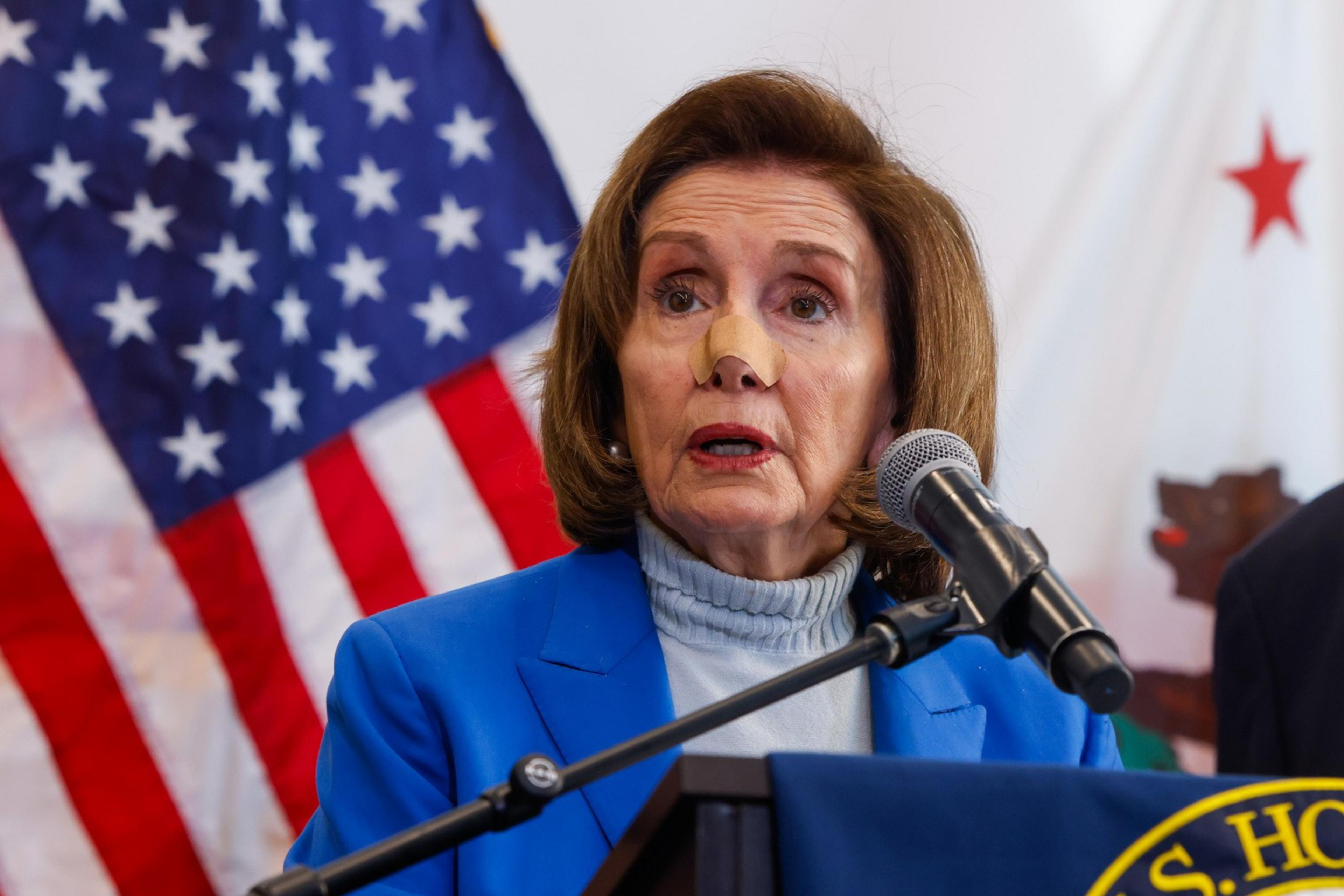 A woman in a blue outfit speaks at a podium with a microphone. An American flag and another person in a suit are in the background. A U.S. House banner is visible.