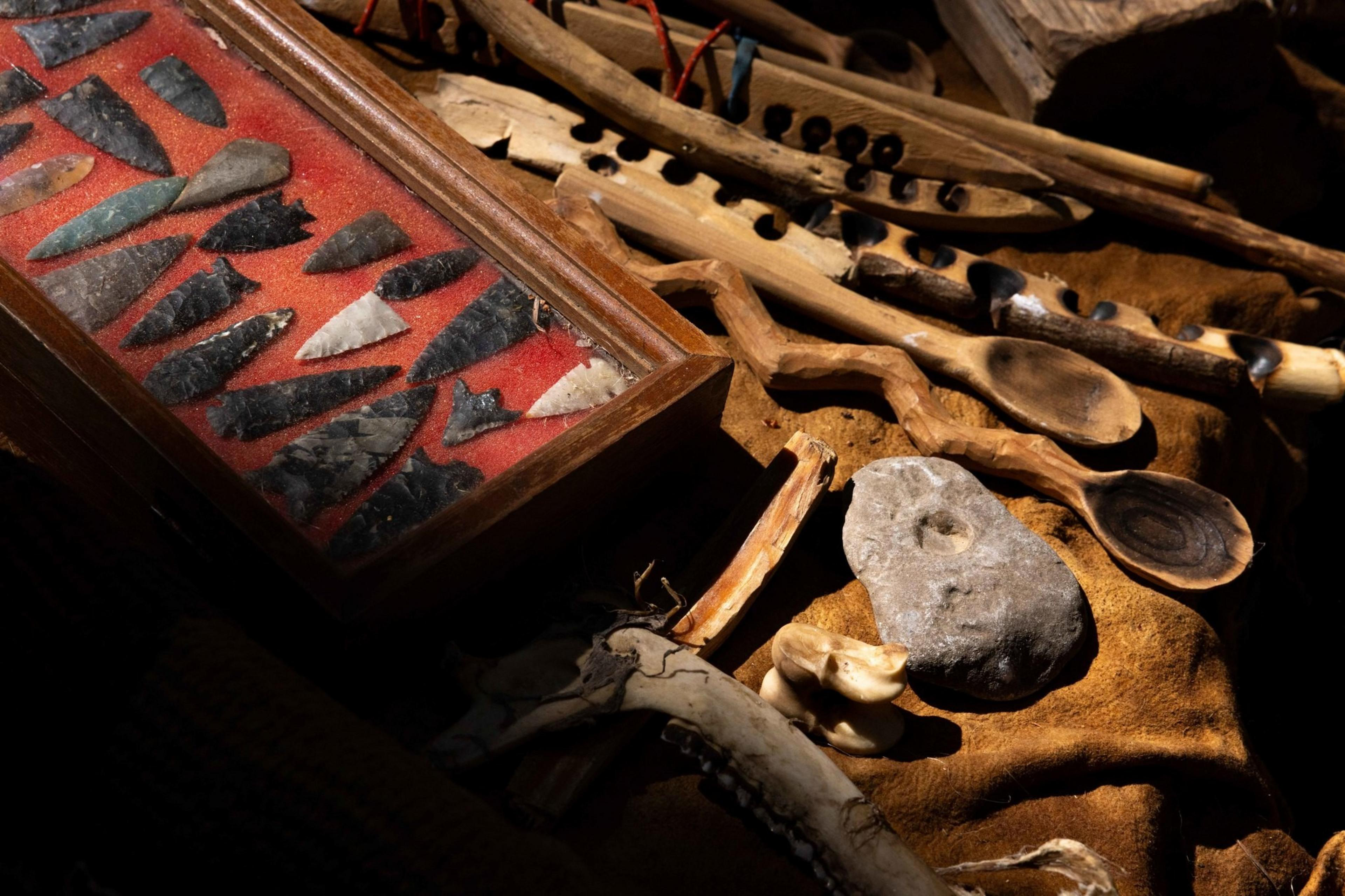 The image shows a collection of stone arrowheads displayed in a glass case, surrounded by carved wooden tools, a stone with a hole, and animal bones on leather.
