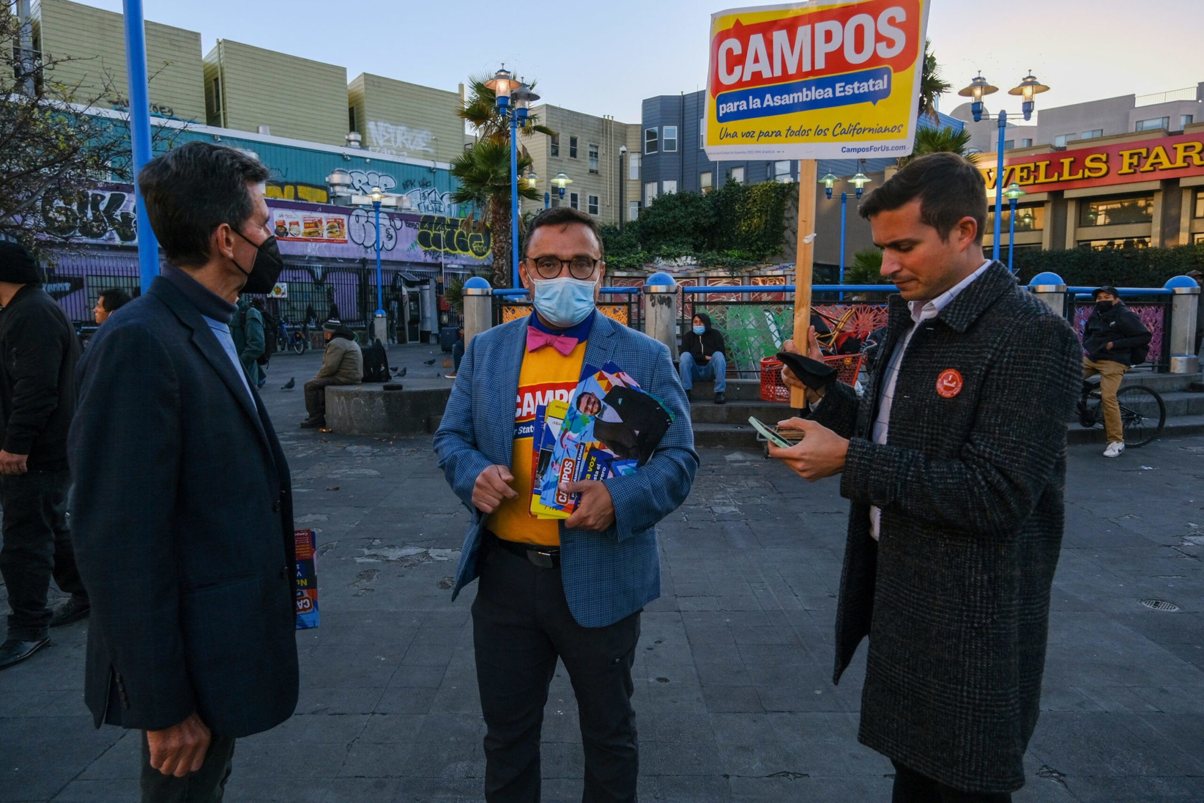 Three men, one holding campaign flyers and a sign for "Campos para la Asamblea Estatal," stand in an outdoor urban area with graffiti and a Wells Fargo building visible.