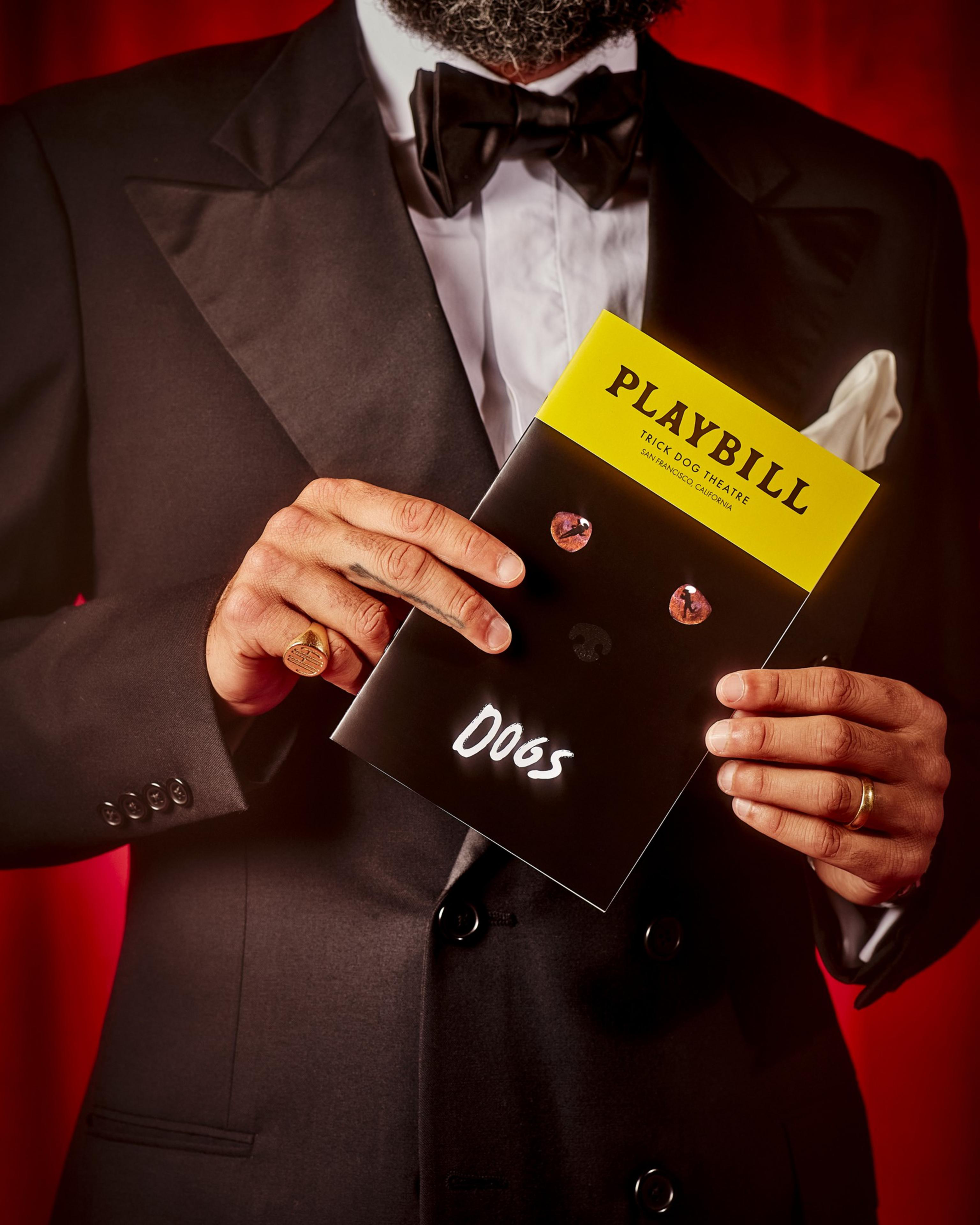 A man in a tuxedo holds a playbill with a yellow top labeled &quot;PLAYBILL&quot; and a black cover with the word &quot;Dogs.&quot; Two round eye-like shapes are on the cover.