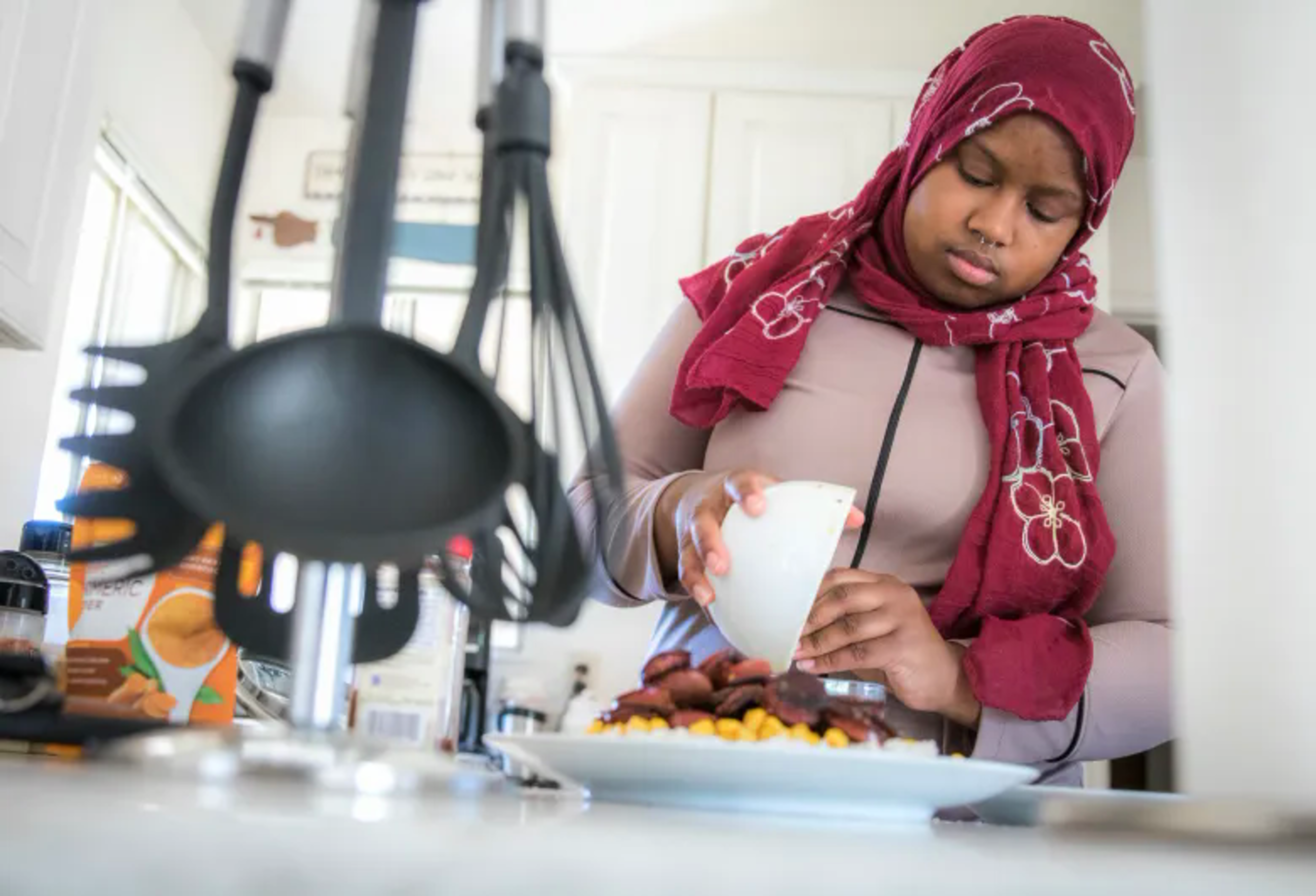 A young woman wearing a red hijab and beige shirt prepares food, pouring from a small bowl onto a plate piled with vegetables and sausage slices in a kitchen.