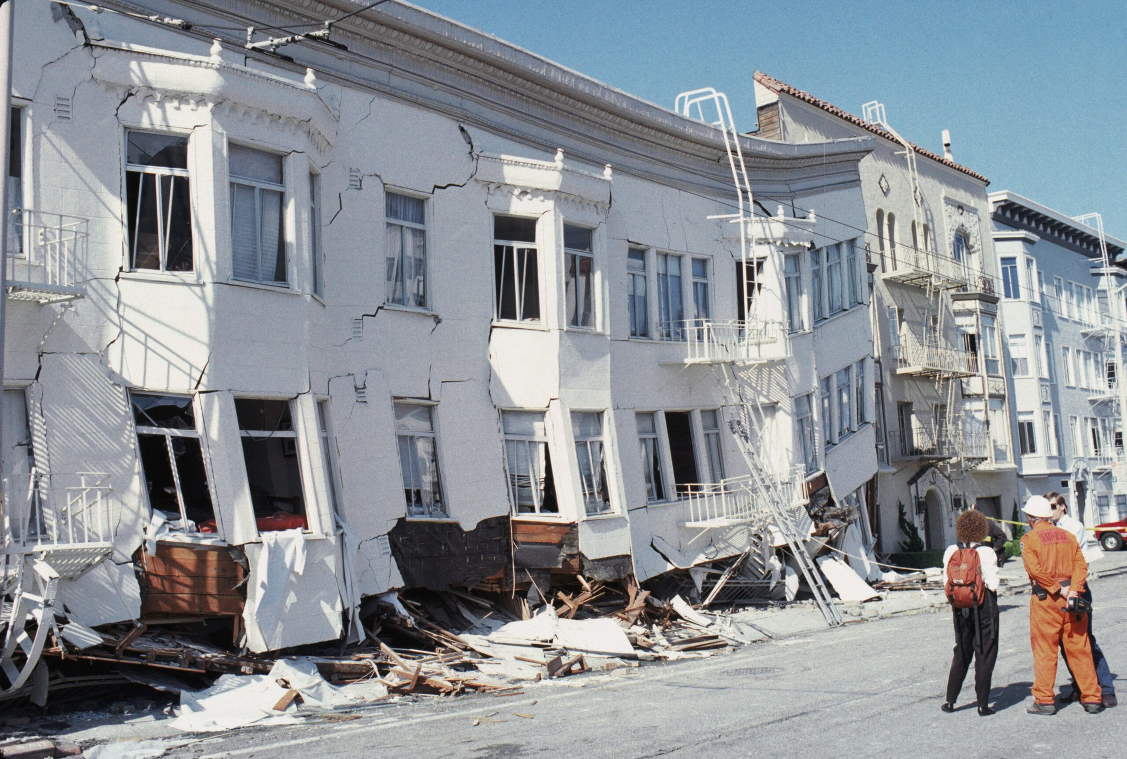 A two-story building is severely damaged, with collapsed walls and fire escapes hanging loosely, while two people in work clothes observe from the street.