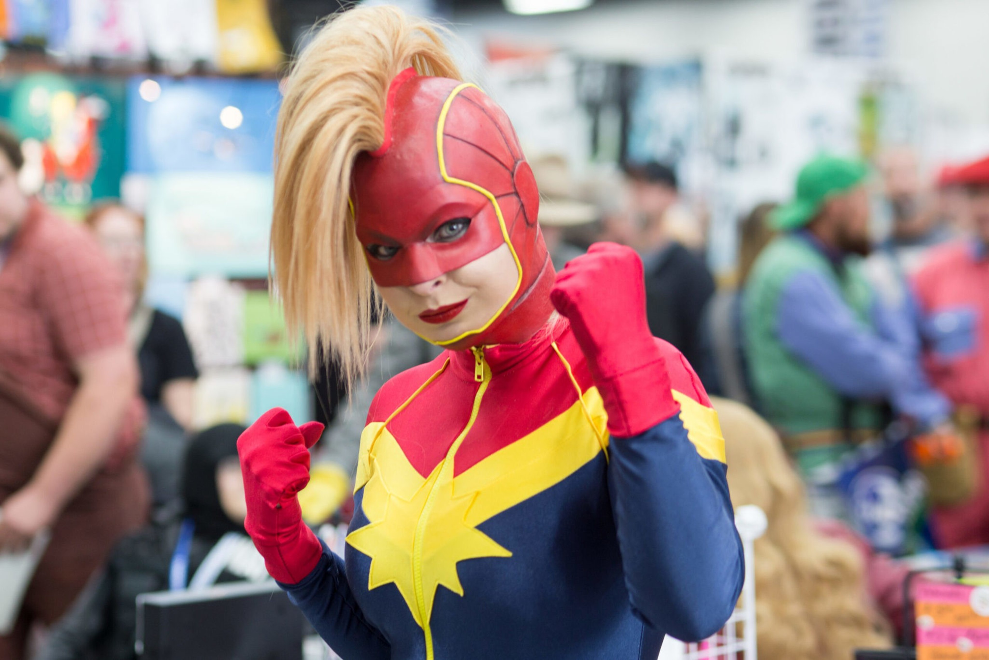 A person dressed as Captain Marvel poses with fists up, wearing a red, blue, and yellow costume with a star emblem and a red mask with blonde hair.