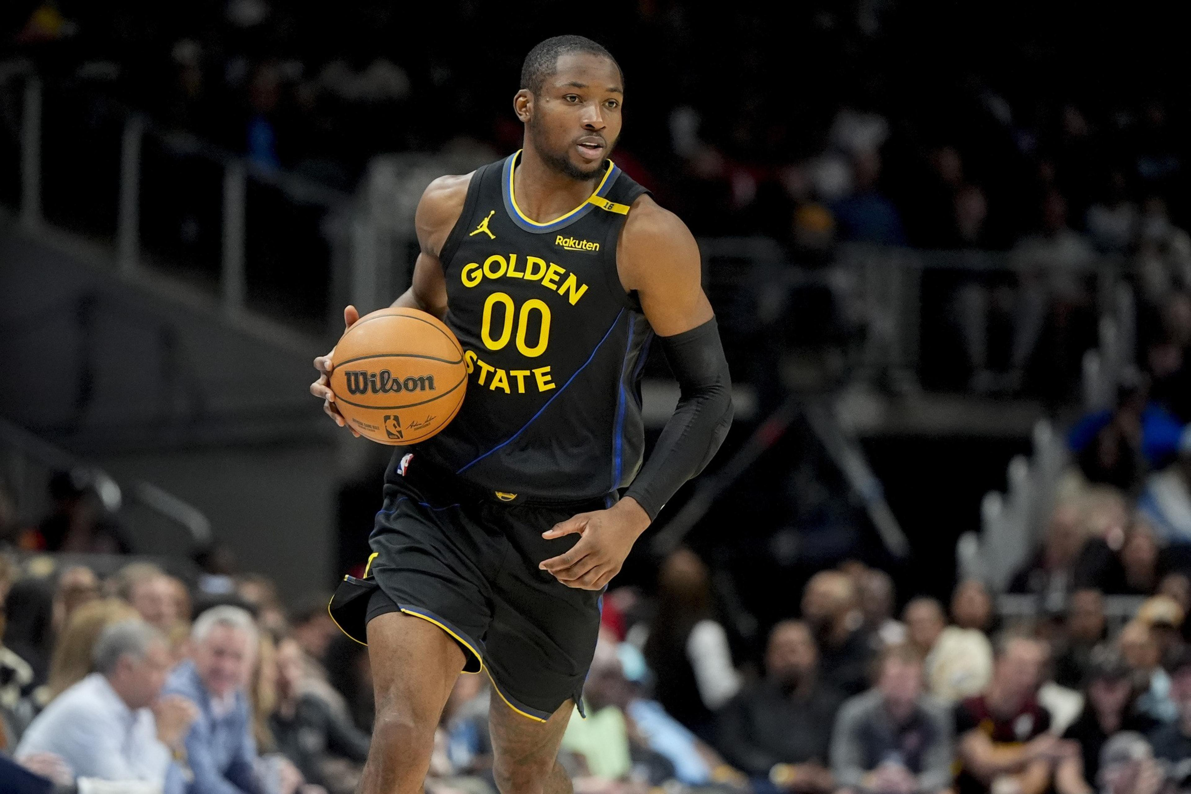 A basketball player wearing a black Golden State jersey dribbles a ball down the court. The audience is blurred in the background.