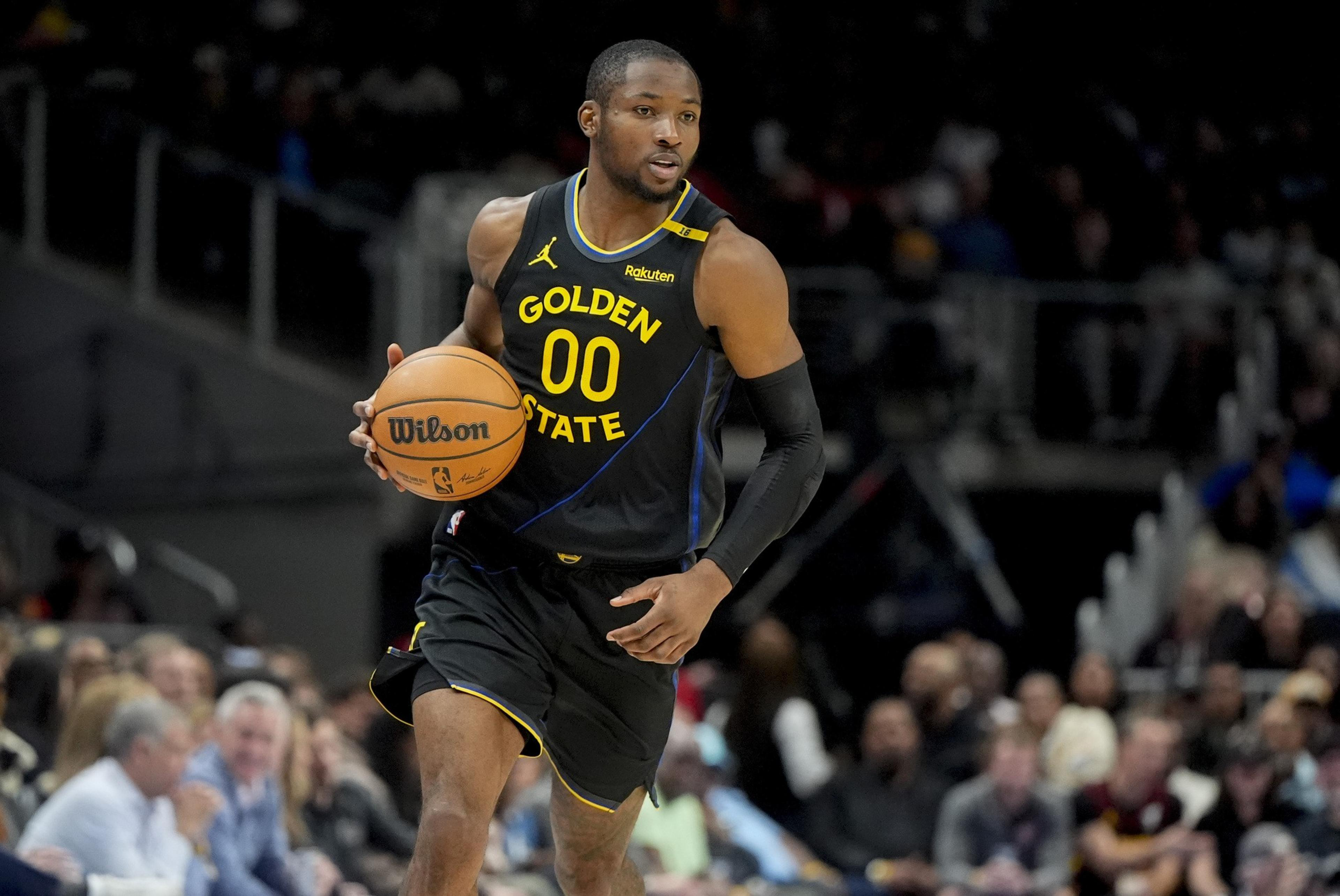 A basketball player wearing a black Golden State jersey dribbles a ball down the court. The audience is blurred in the background.