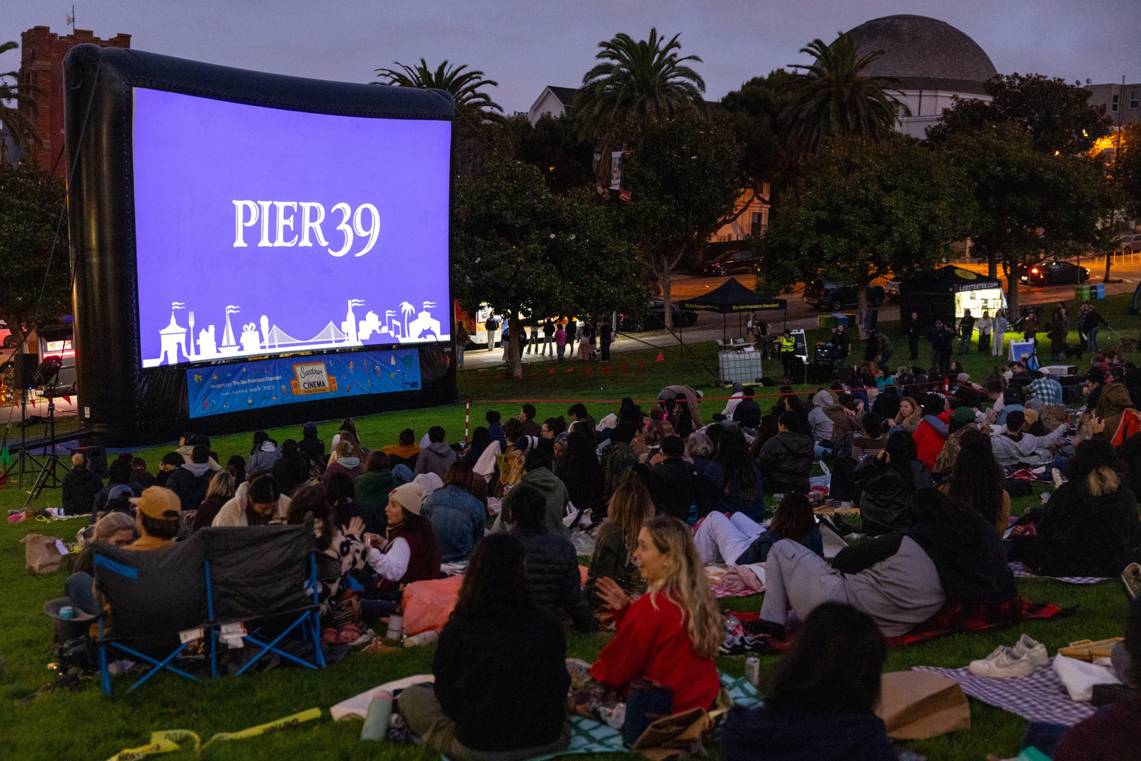 People sit on blankets and lawn chairs on a grassy hill in the evening, watching a large outdoor movie screen that reads “PIER 39.”