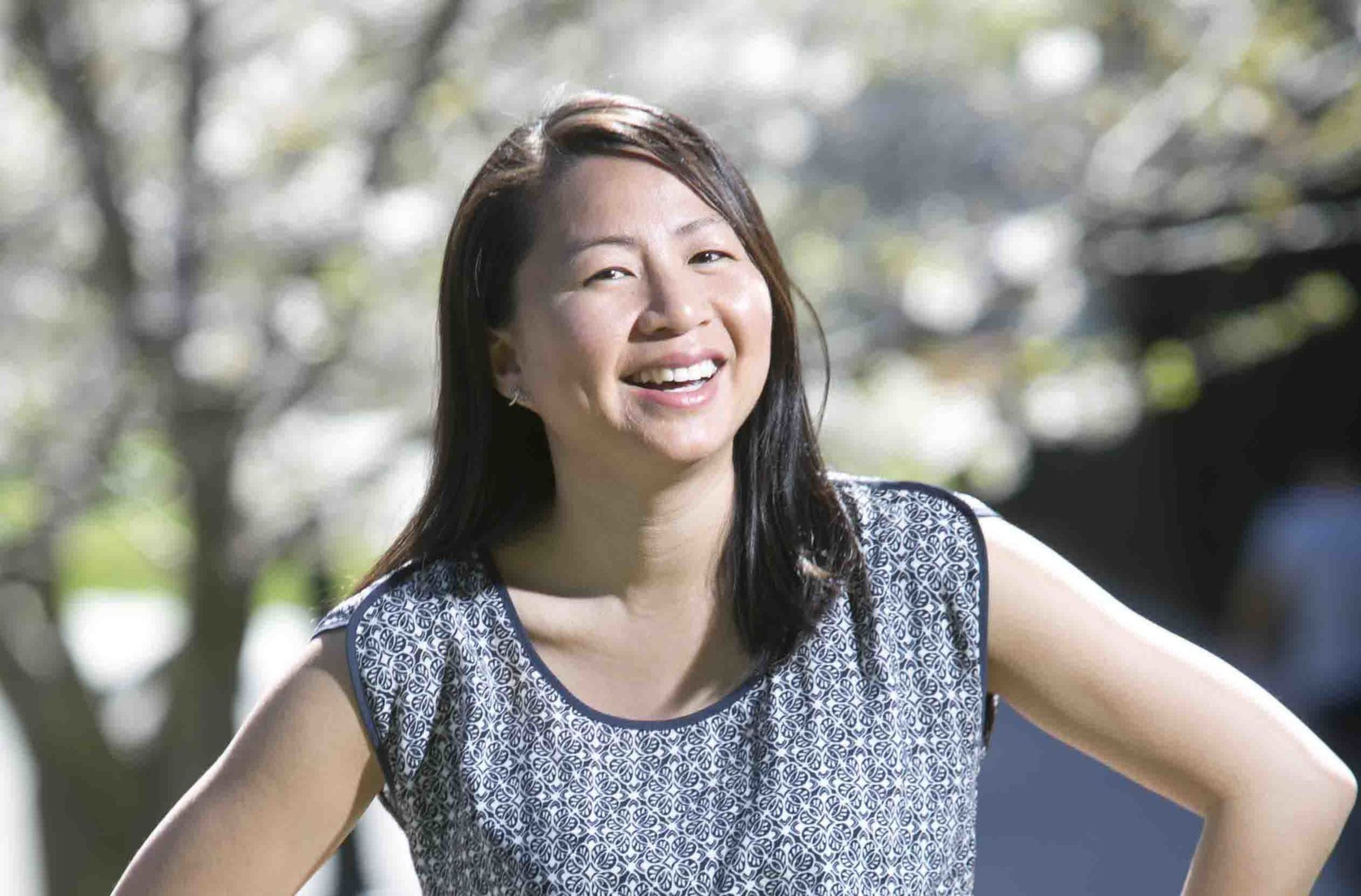 A woman with long dark hair is smiling broadly while standing with her hands on her hips, wearing a sleeveless patterned top outdoors.