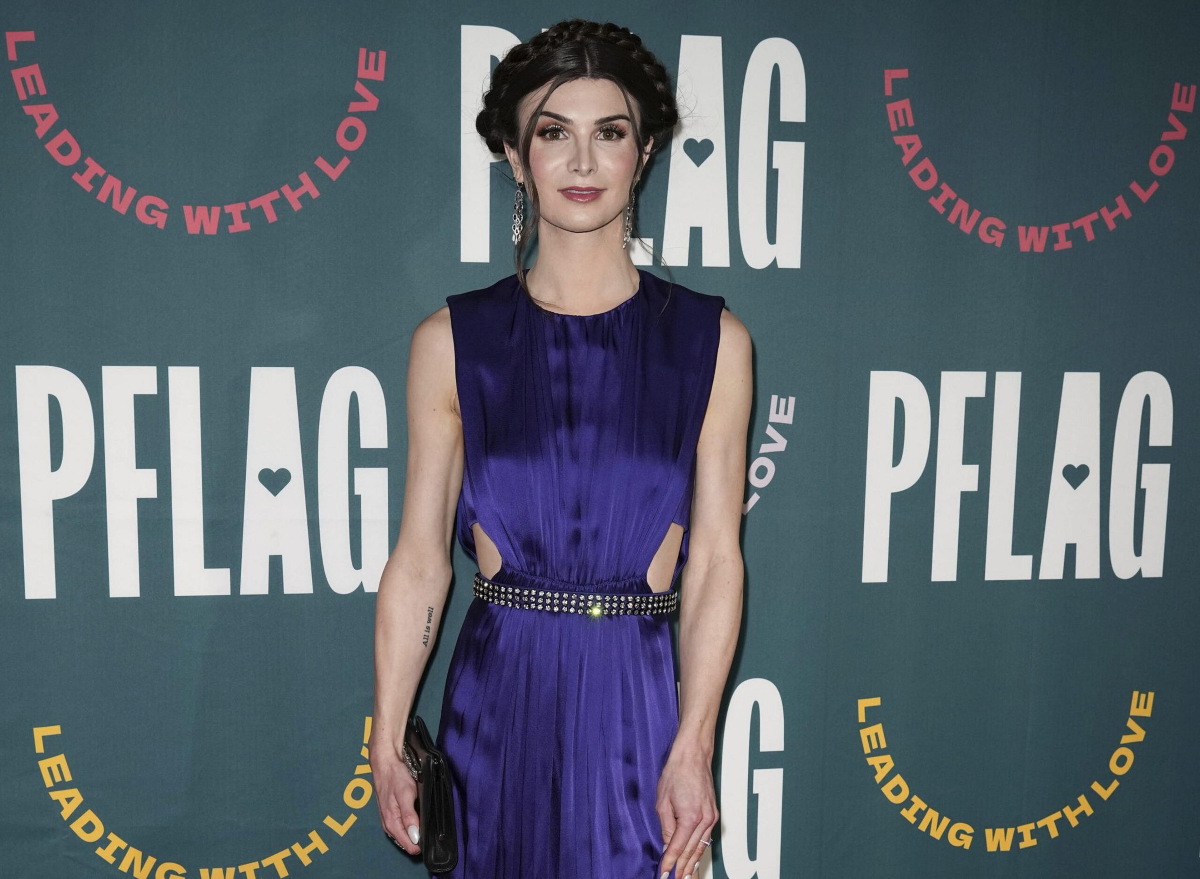 A woman with braided hair wears a sleeveless, purple satin dress with cutouts and a studded belt, standing in front of a "PFLAG" backdrop.