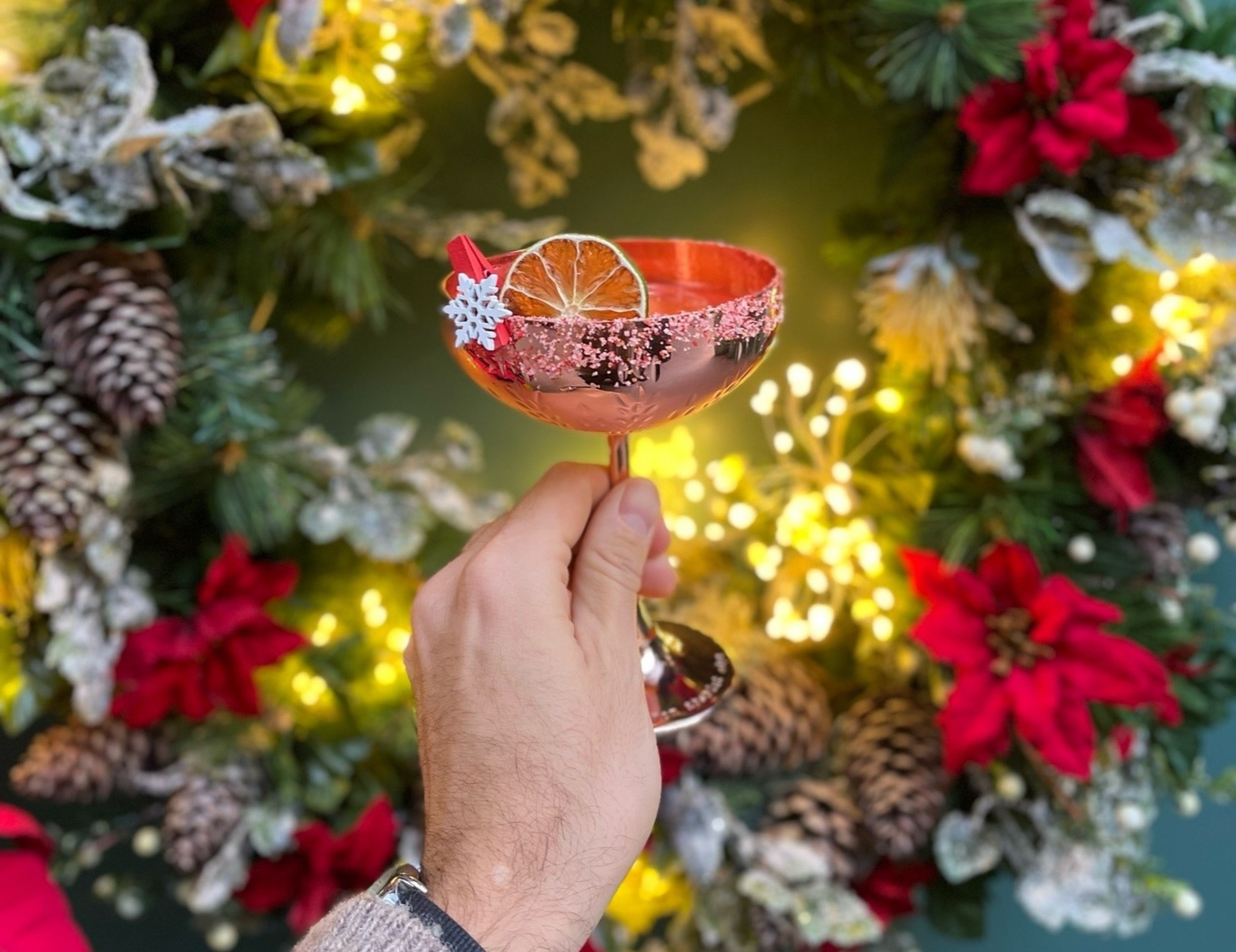 A cocktail is held up in a brass coupe martini glass against a wreathed background.