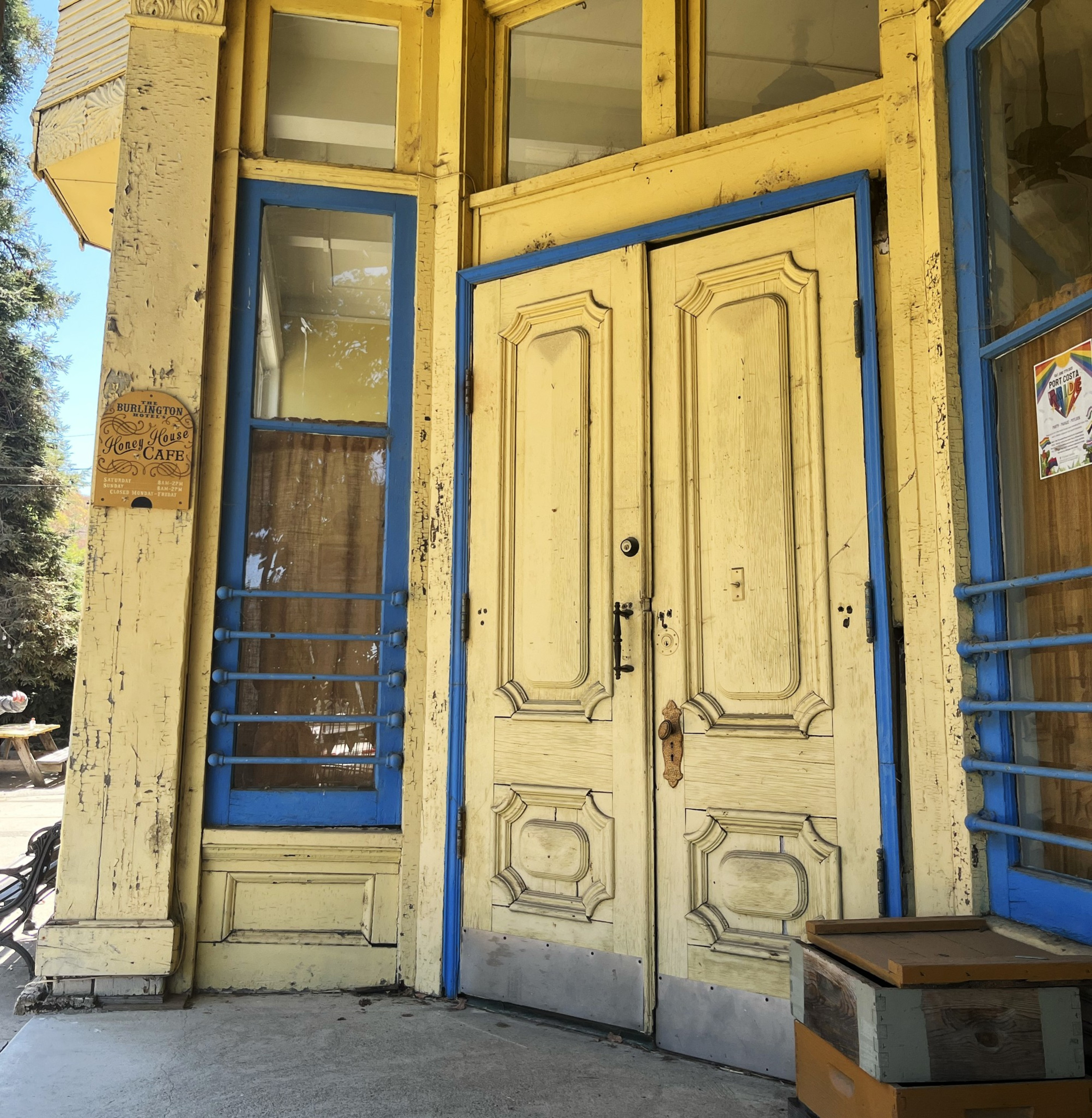Yellow double doors with ornate panels and blue trim stand closed on a weathered wooden café building with glass windows and a small wooden box nearby.