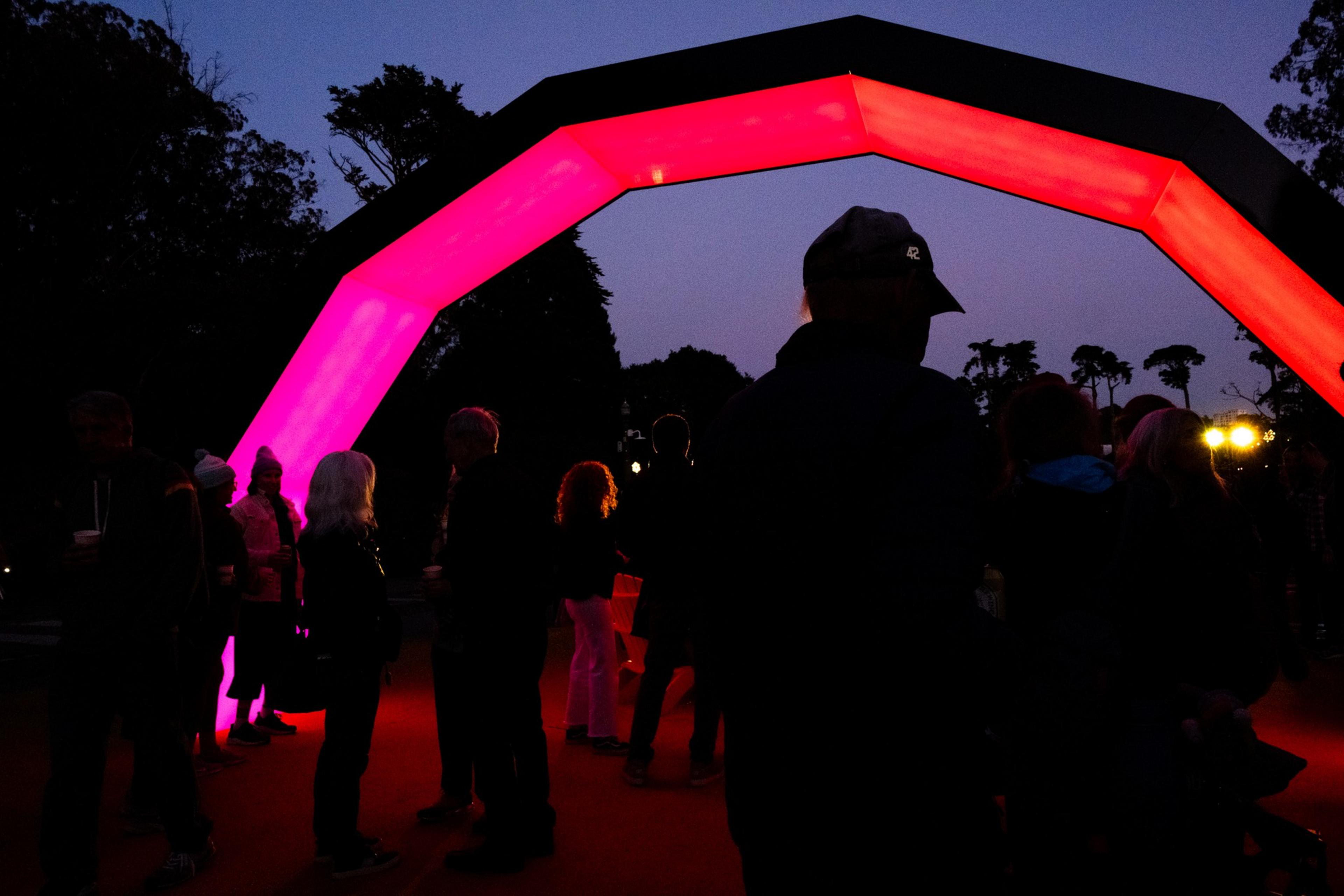 A group of people stand under a glowing pink archway in a dark outdoor setting, silhouetted against the evening sky with trees in the background.