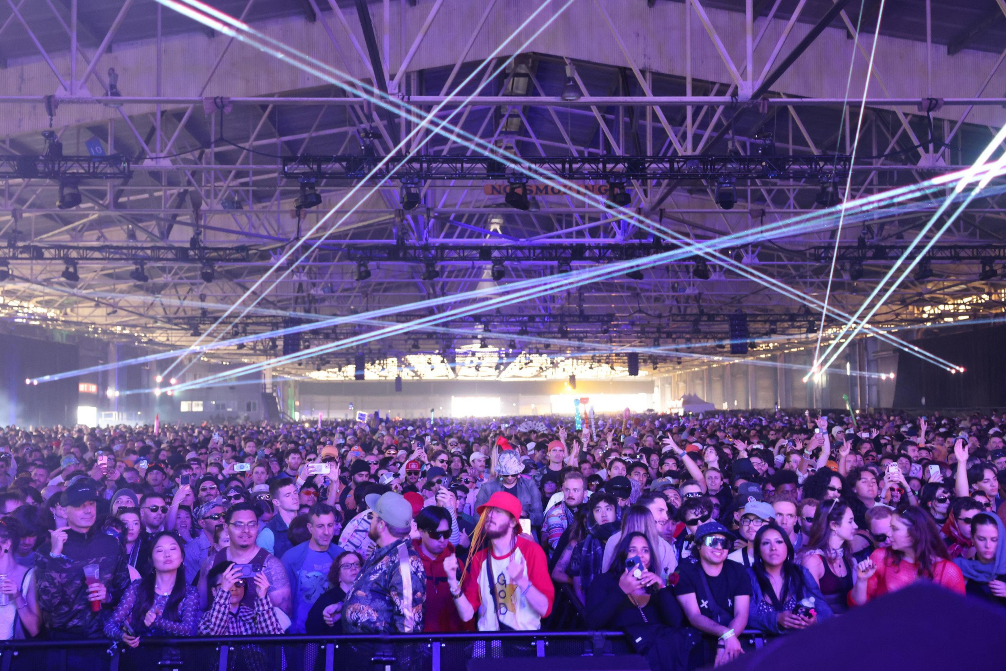 A large crowd of people is gathered indoors at an event with colorful lasers crisscrossing the ceiling, creating a vibrant and energetic atmosphere.