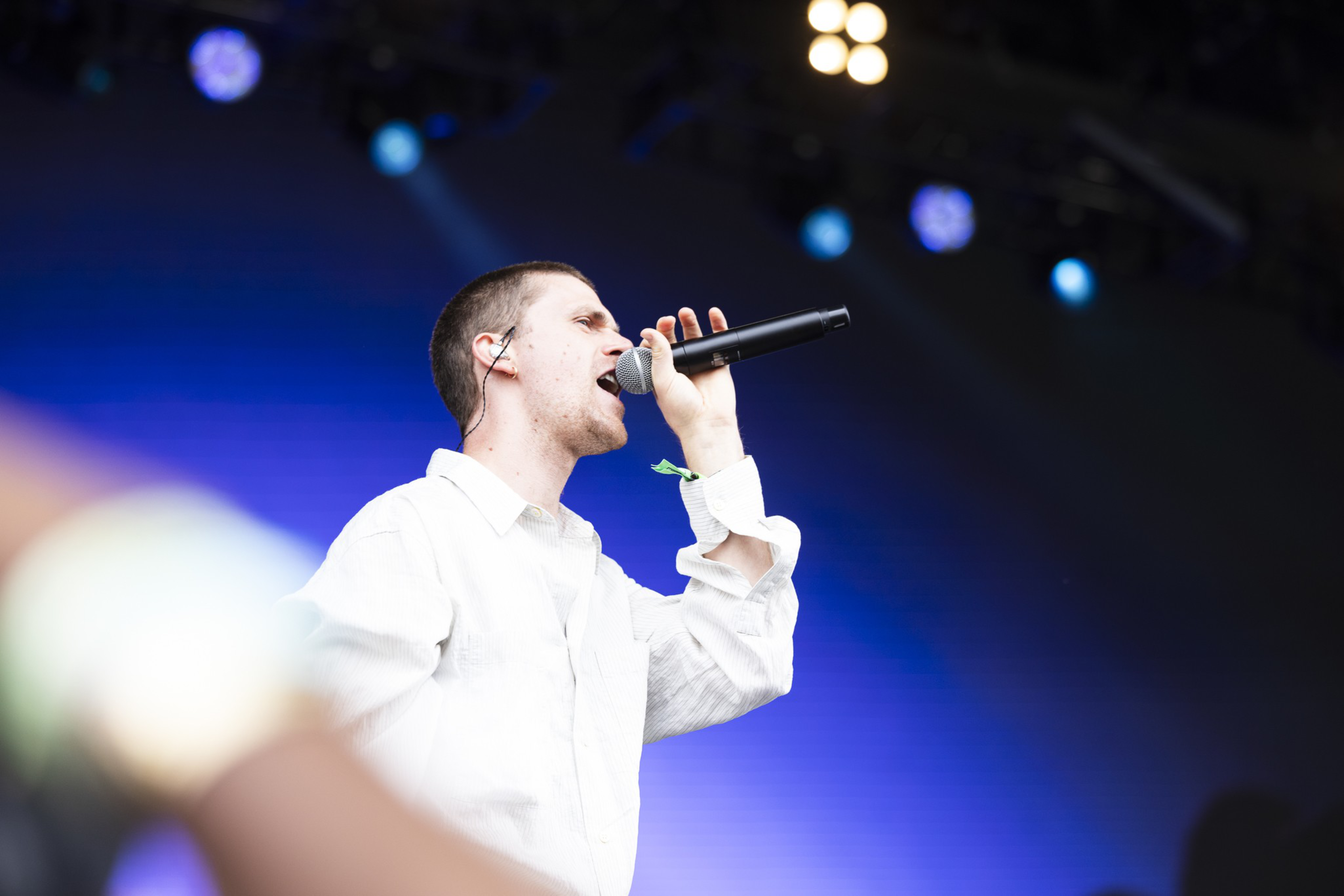 A young man wearing a white shirt is singing into a microphone on stage, with stage lights and a blue background behind him.