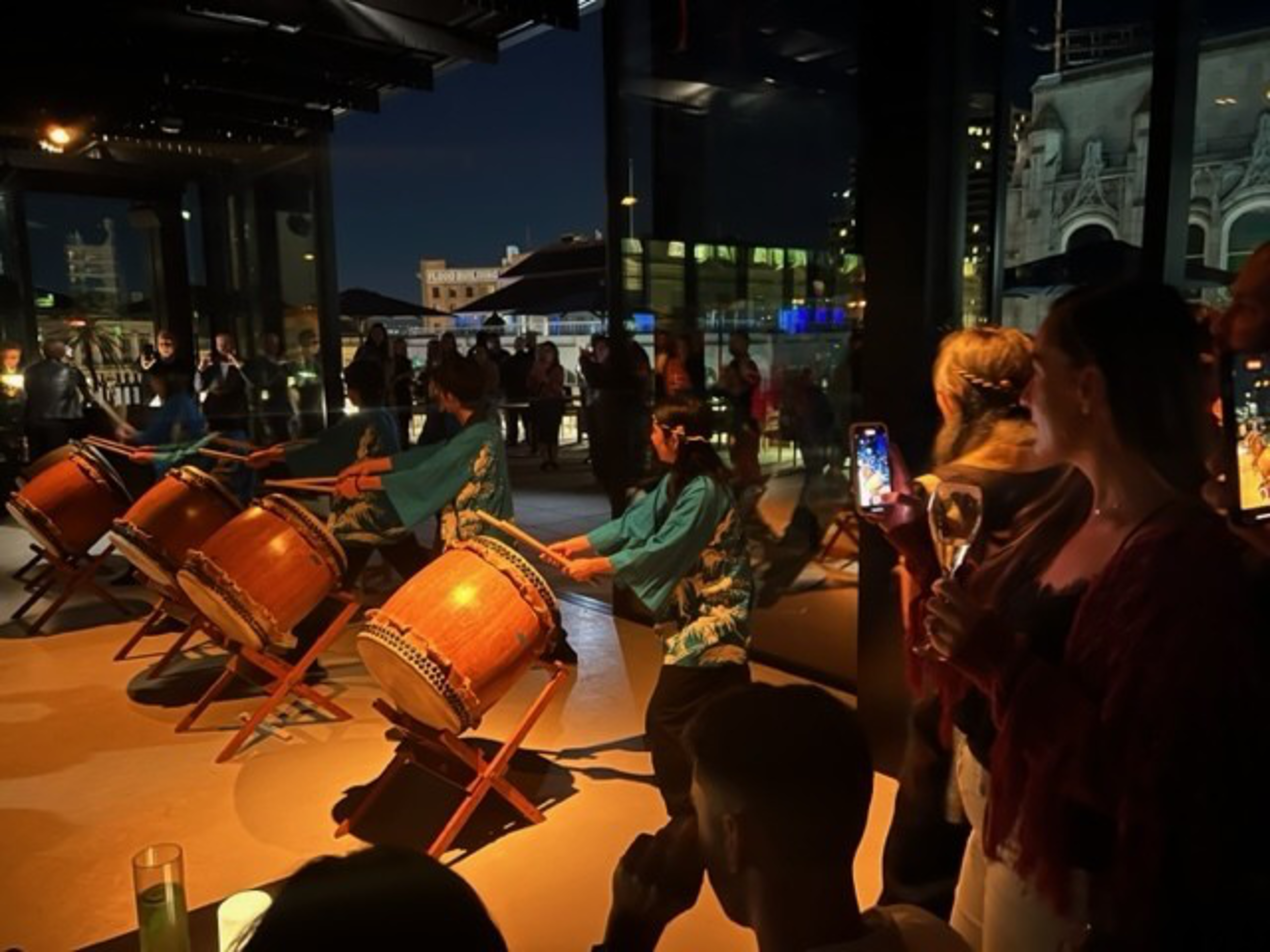 Five drummers wearing blue and white traditional outfits perform on large wooden drums while a crowd watches, some filming the event on phones.