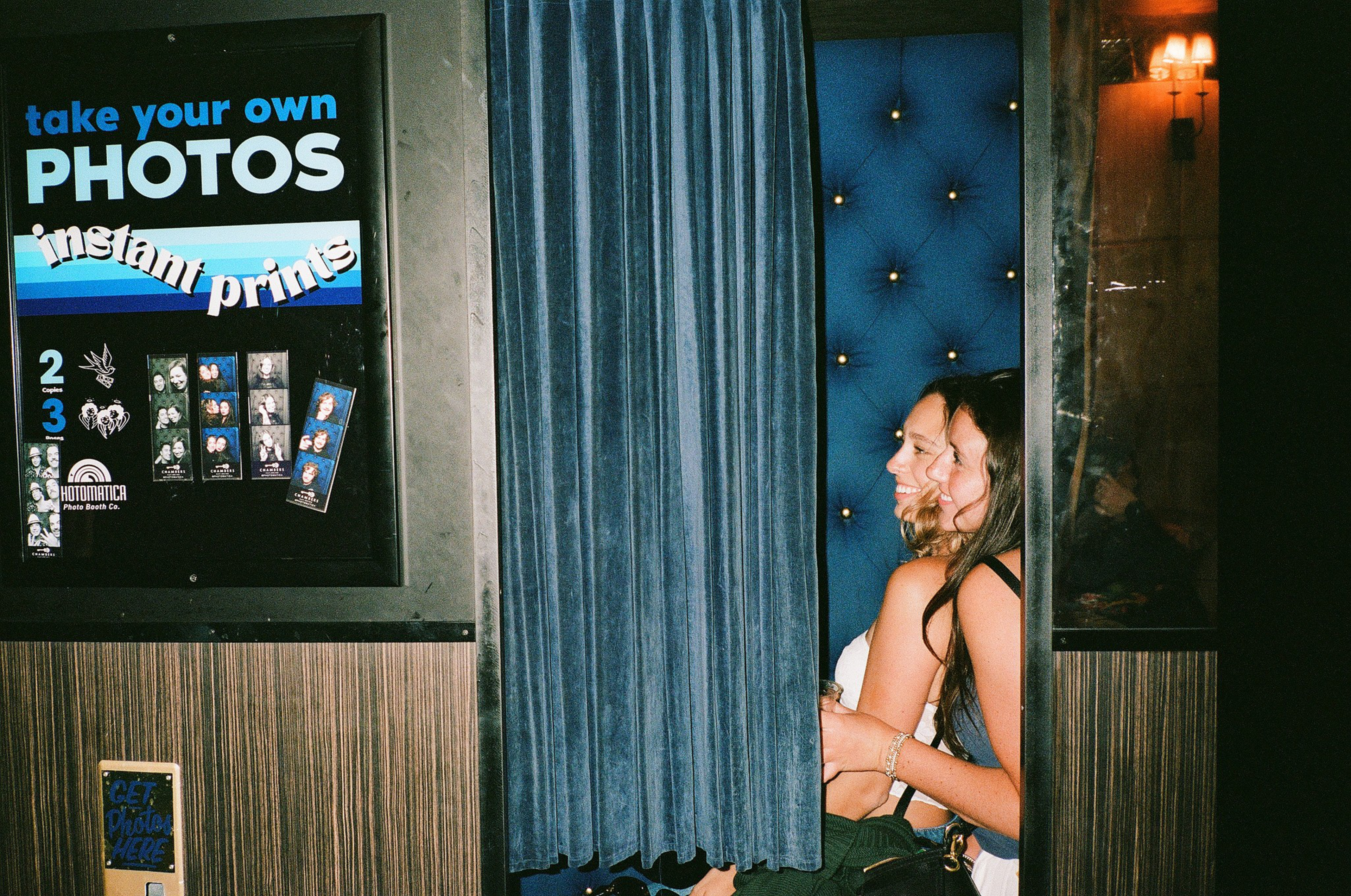 Two women smile inside a photo booth with a blue velvet curtain and padded blue back wall, next to a sign advertising instant photo prints.