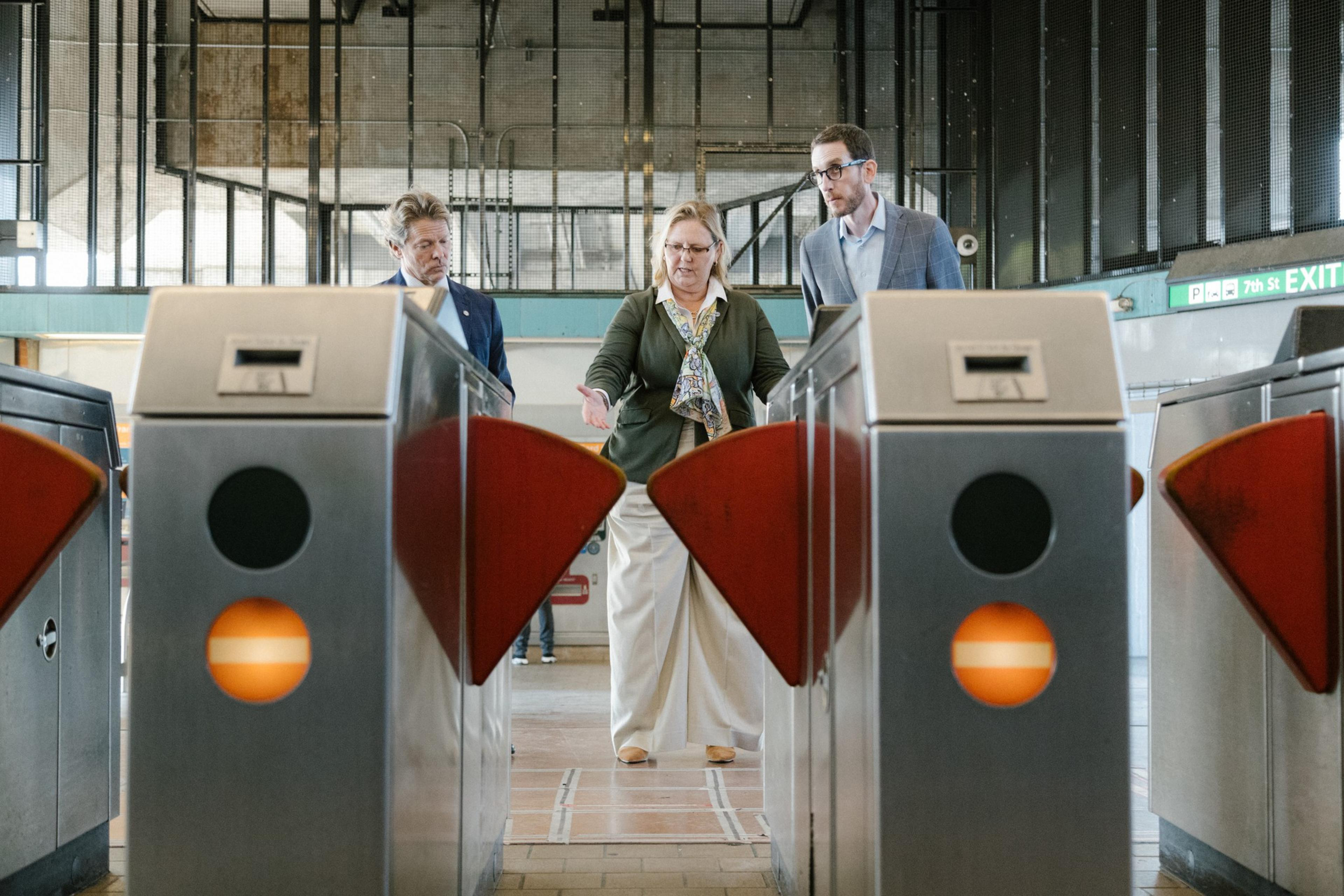Three people stand behind turnstiles at a transit station, seemingly discussing or observing the gates, with an exit sign visible in the background.