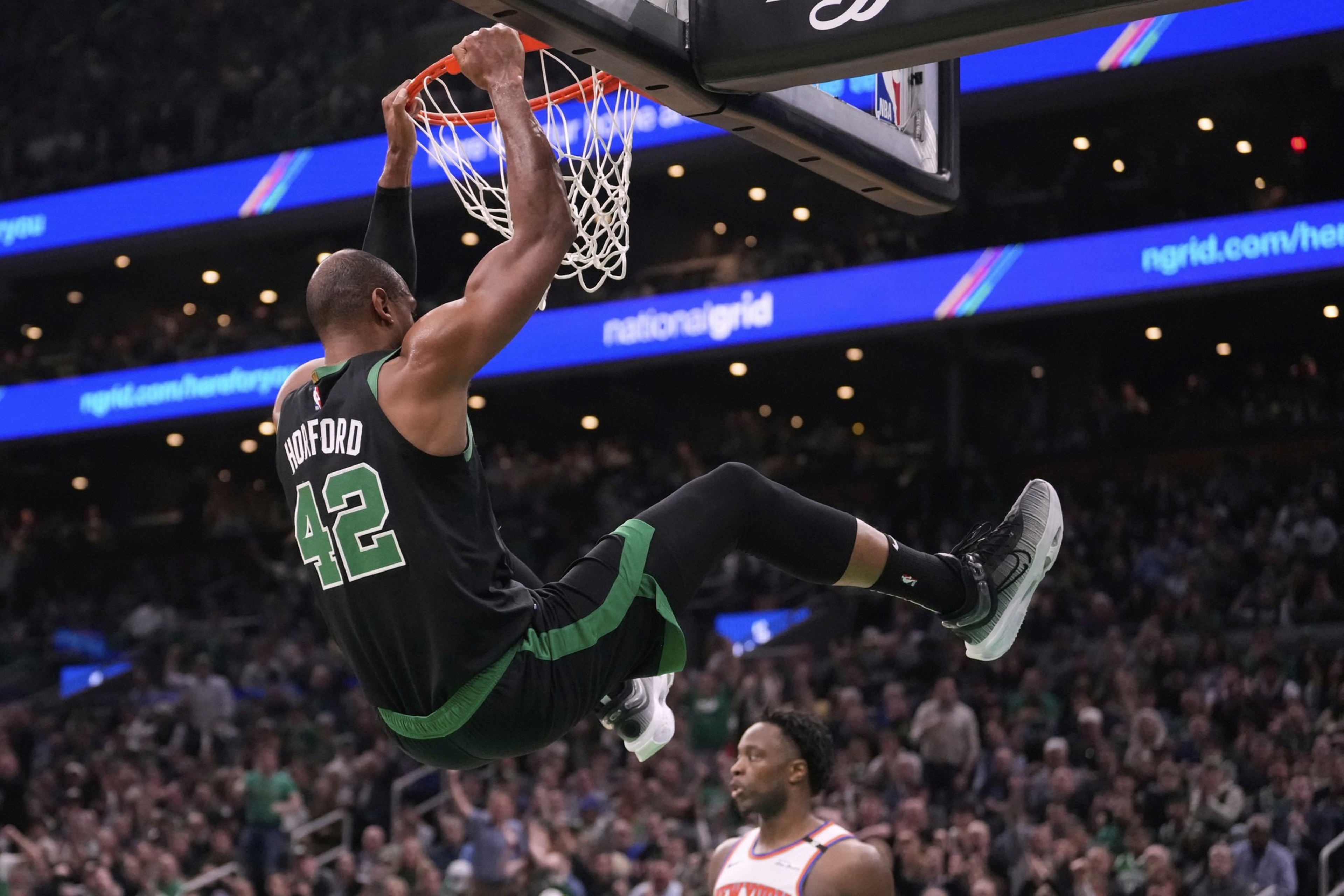 A basketball player in a green jersey labeled &quot;42&quot; hangs from the rim after dunking. Another player in a white jersey watches, with a crowded arena in the background.