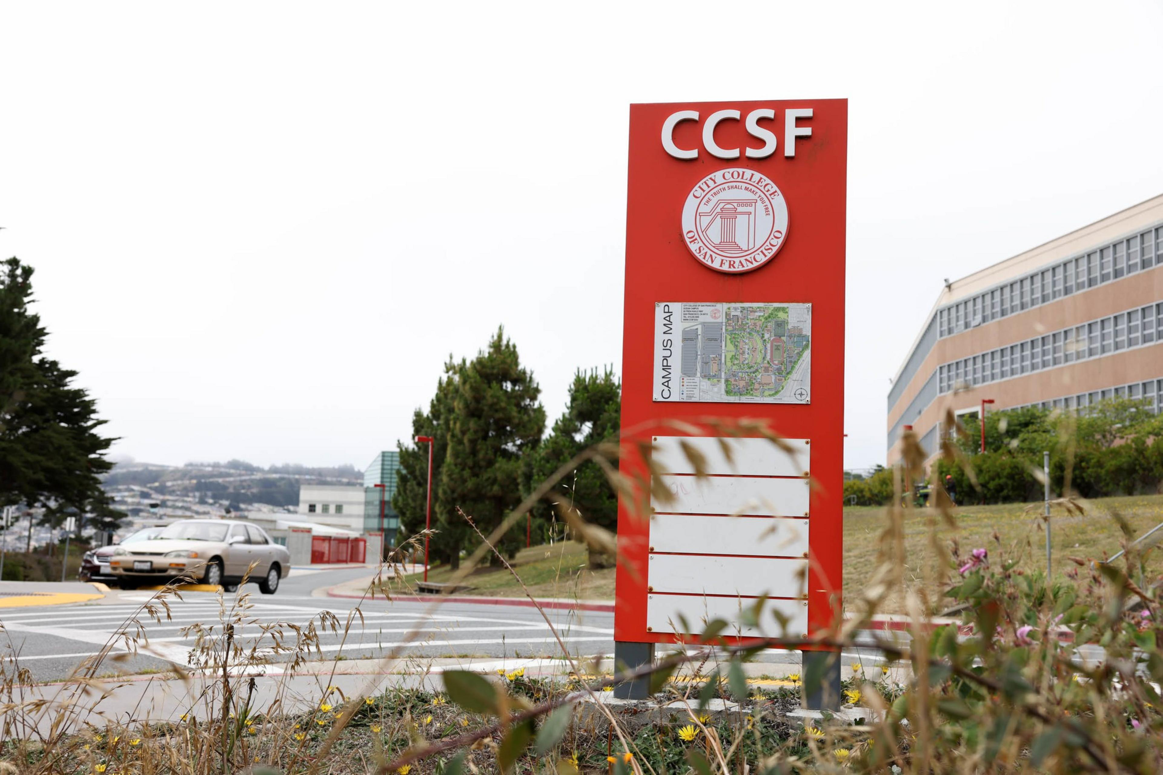 A red sign for City College of San Francisco with a campus map stands near a road, surrounded by grass and plants, with cars and buildings in the background.