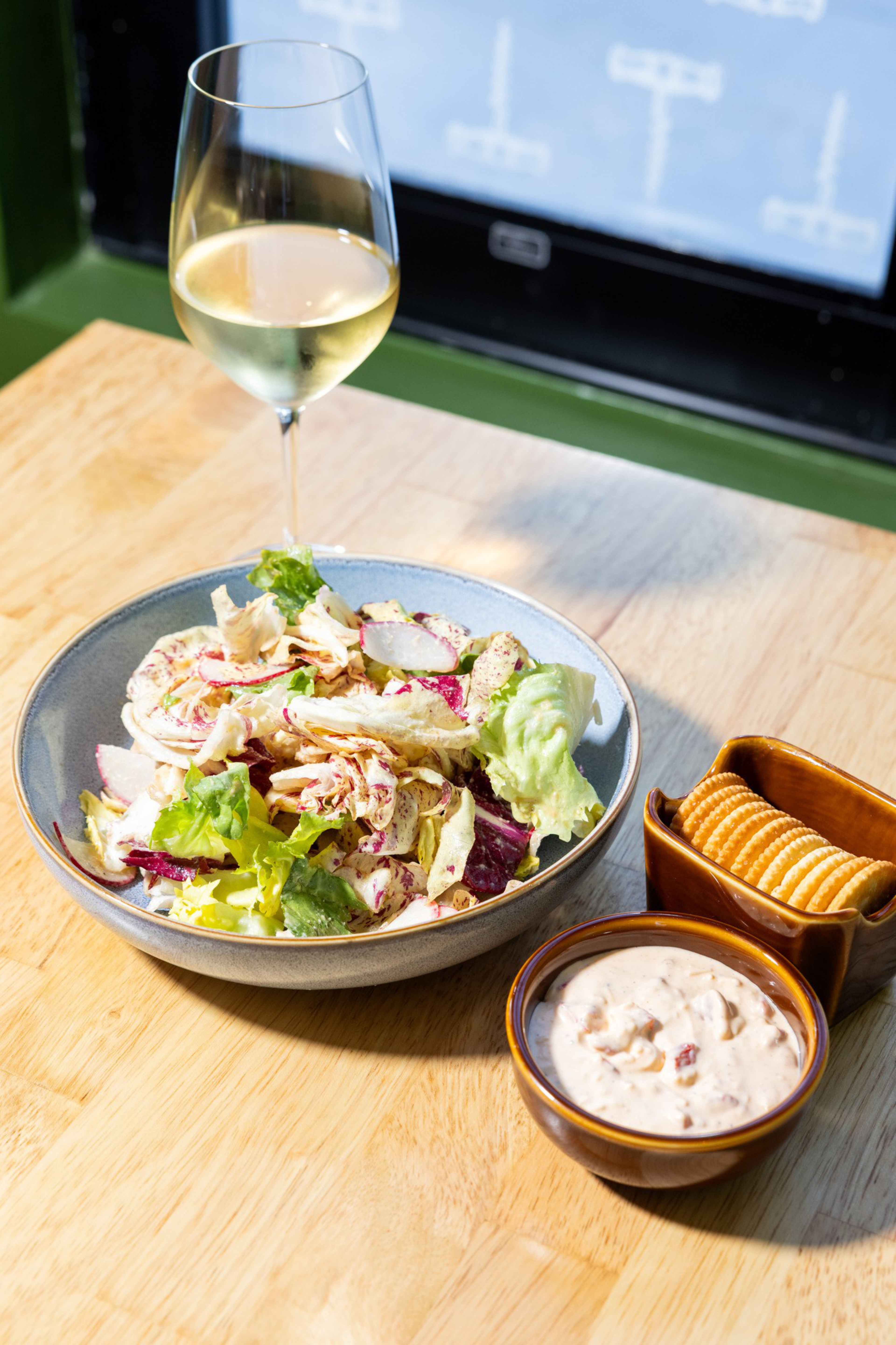 A plate of mixed salad with a variety of greens sits on a wooden table. Beside it are a bowl of creamy dip and a dish of round crackers. A glass of white wine is also present.
