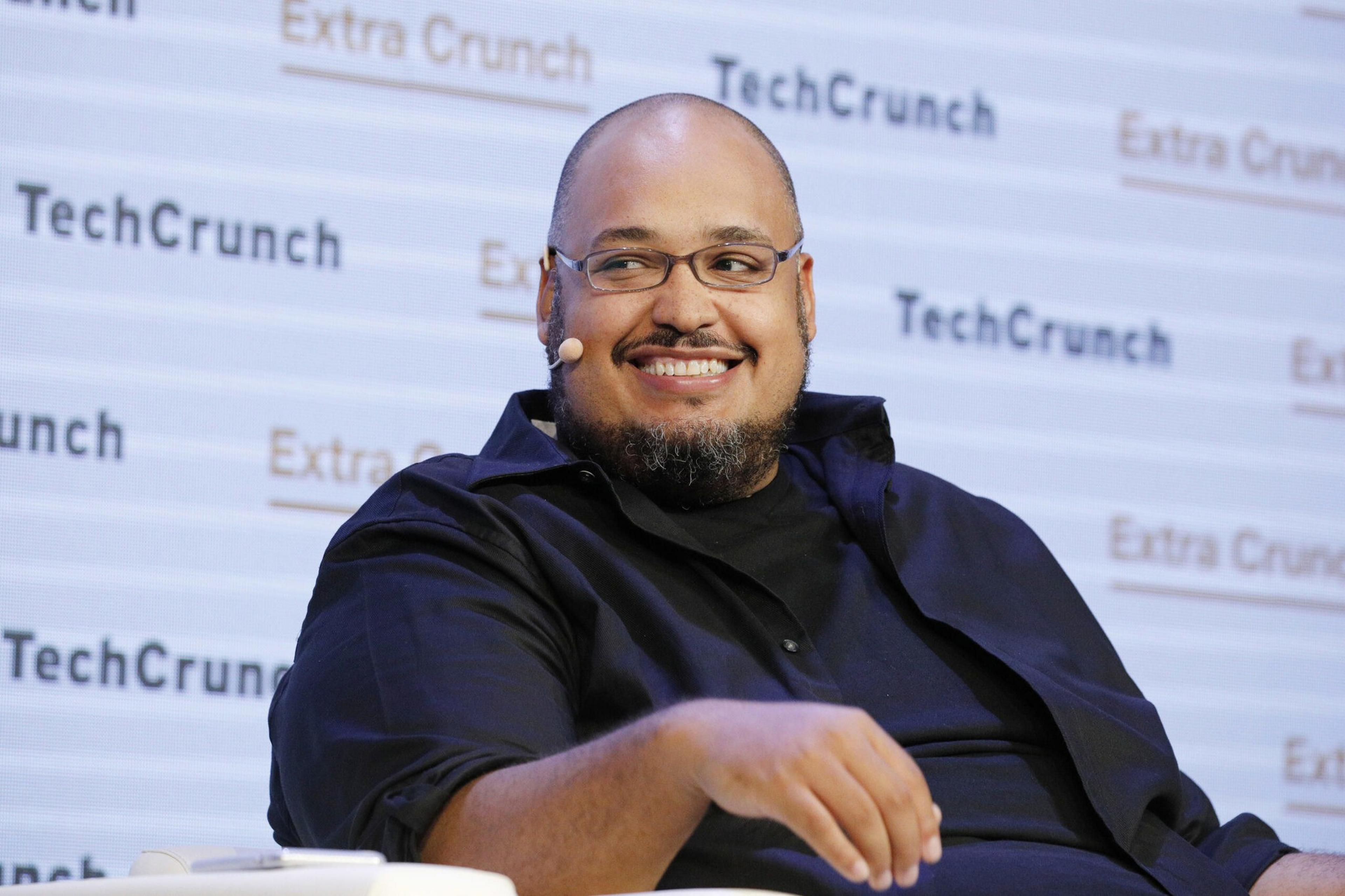 A smiling man with glasses, a beard, and a bald head is sitting and wearing a black shirt with a microphone on his cheek.