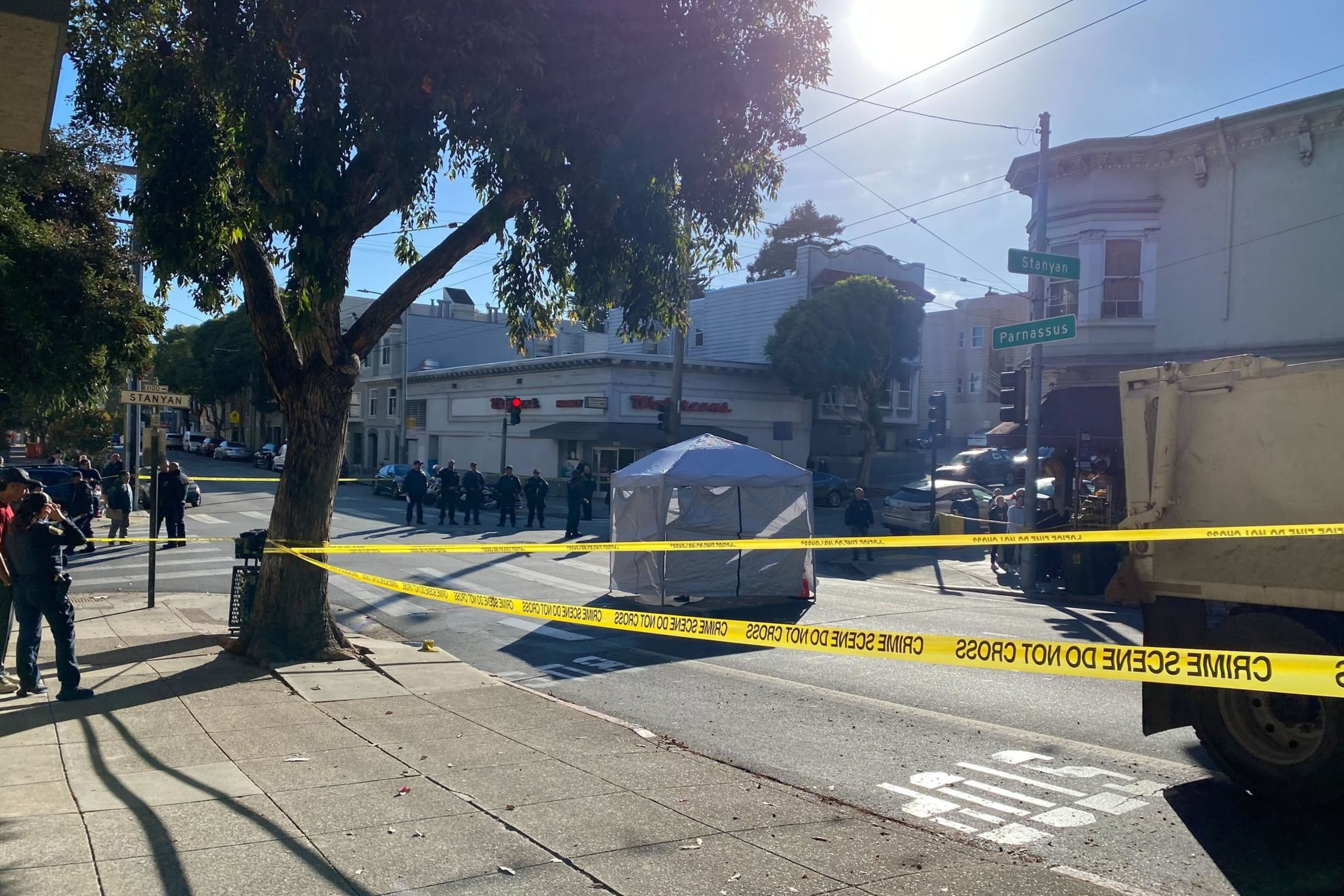 A police scene is cordoned off with yellow tape at the corner of Stanyan and Parnassus streets. Officers and a small tent are present under a clear, sunny sky.