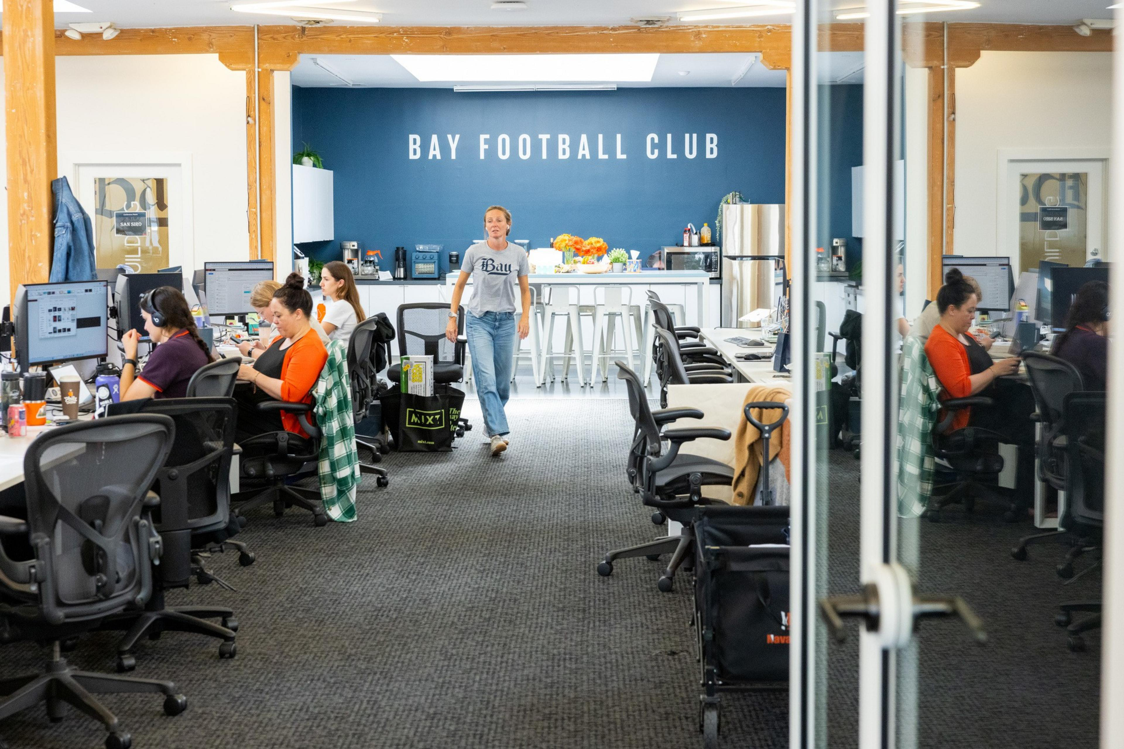 A woman walks toward the camera in an open office with several people working at desks, a kitchen area, and "Bay Football Club" on a blue wall.