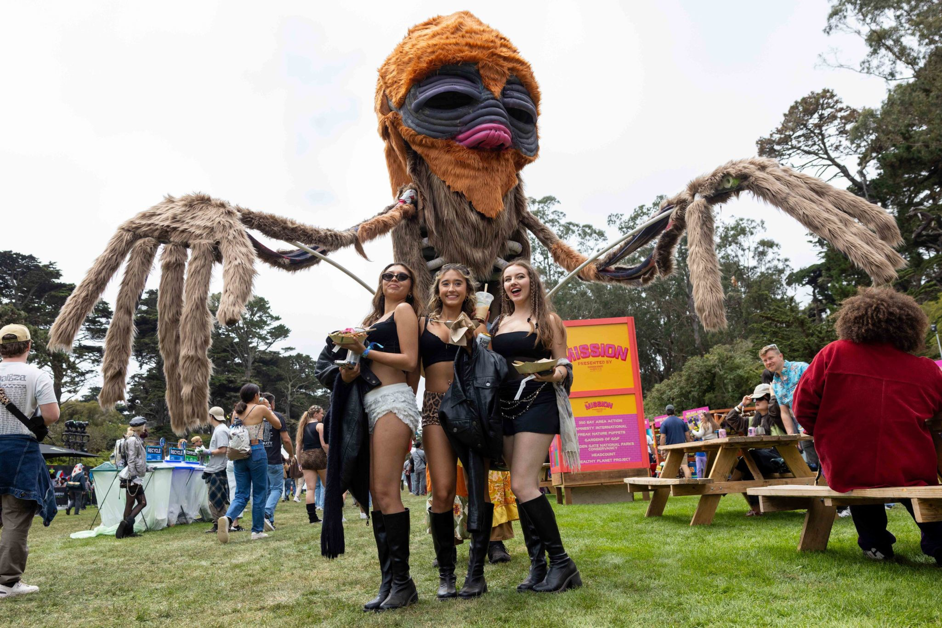 Three women in festival attire pose happily in front of a large spider-like puppet with long furry legs and a colorful face at an outdoor event.