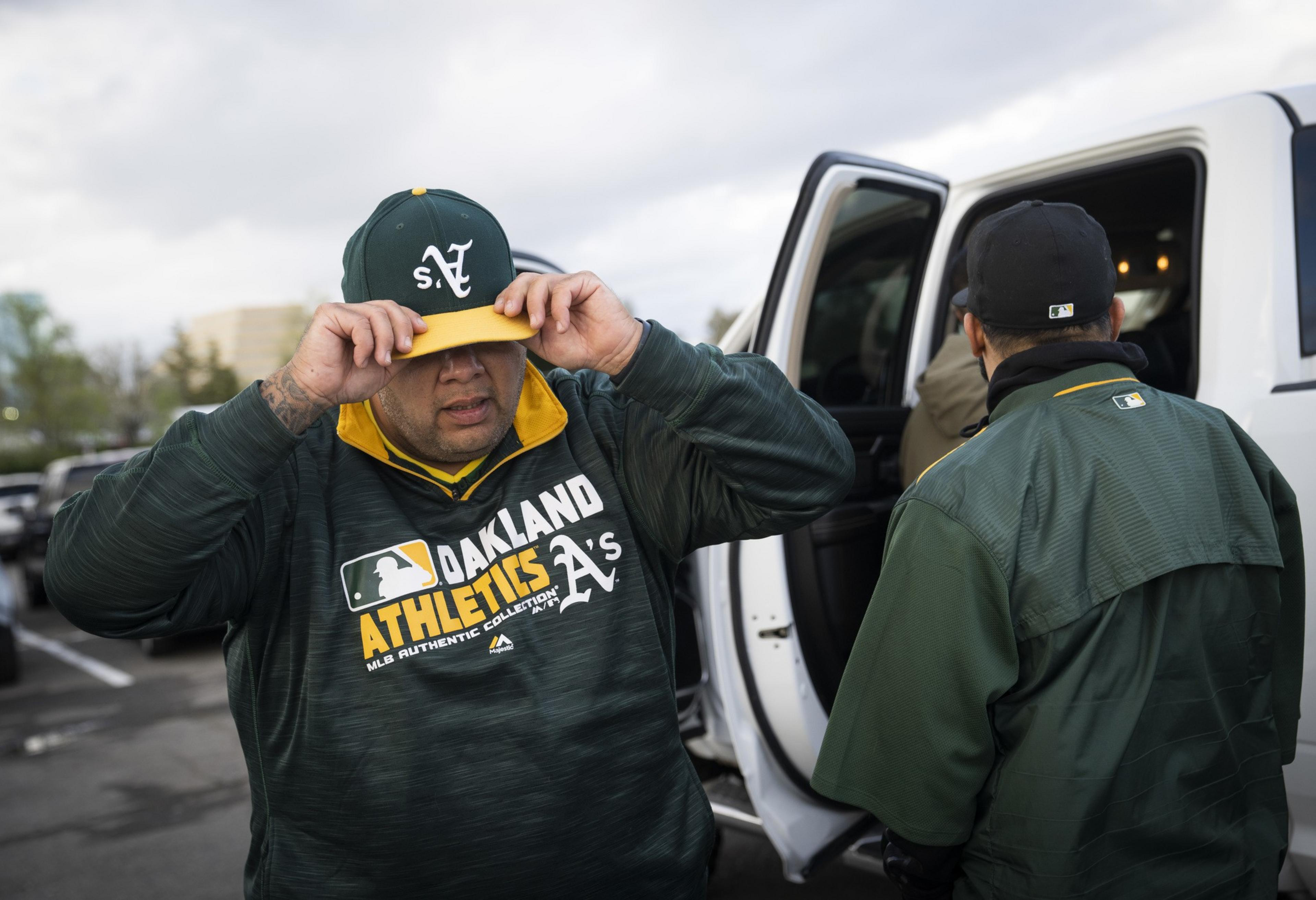 A person adjusts a cap labeled &quot;SV&quot; and wears an Oakland Athletics sweatshirt, standing next to a white vehicle with another person nearby.