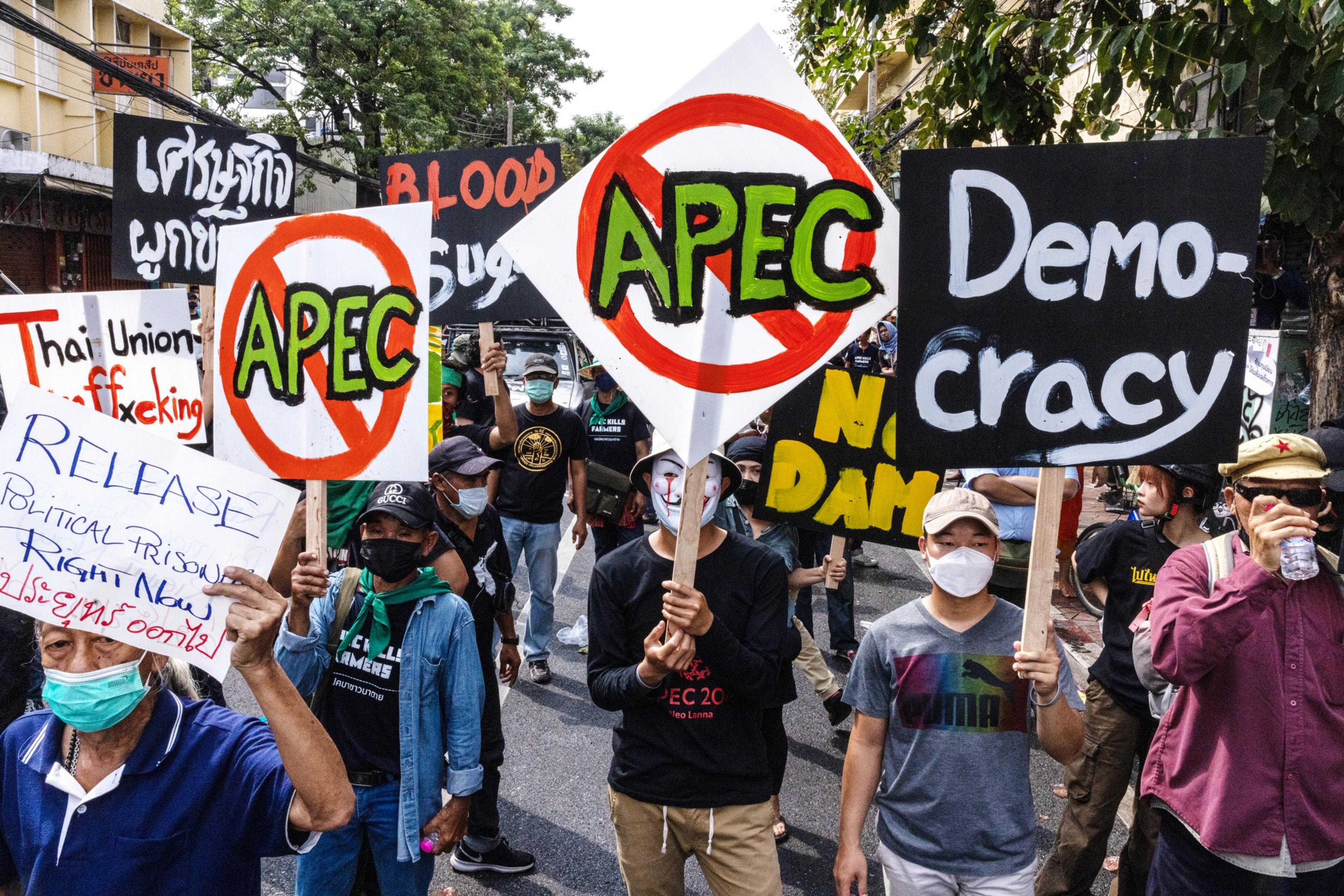 Dozens of people wearing COVID face mask stand closely together in the middle of a street in Bangkok, Thailand, during an Anti-APEC protest. They are holding signs that read "Democracy" "Release Political Prisoners Right Now" and the acronym "APEC" with a