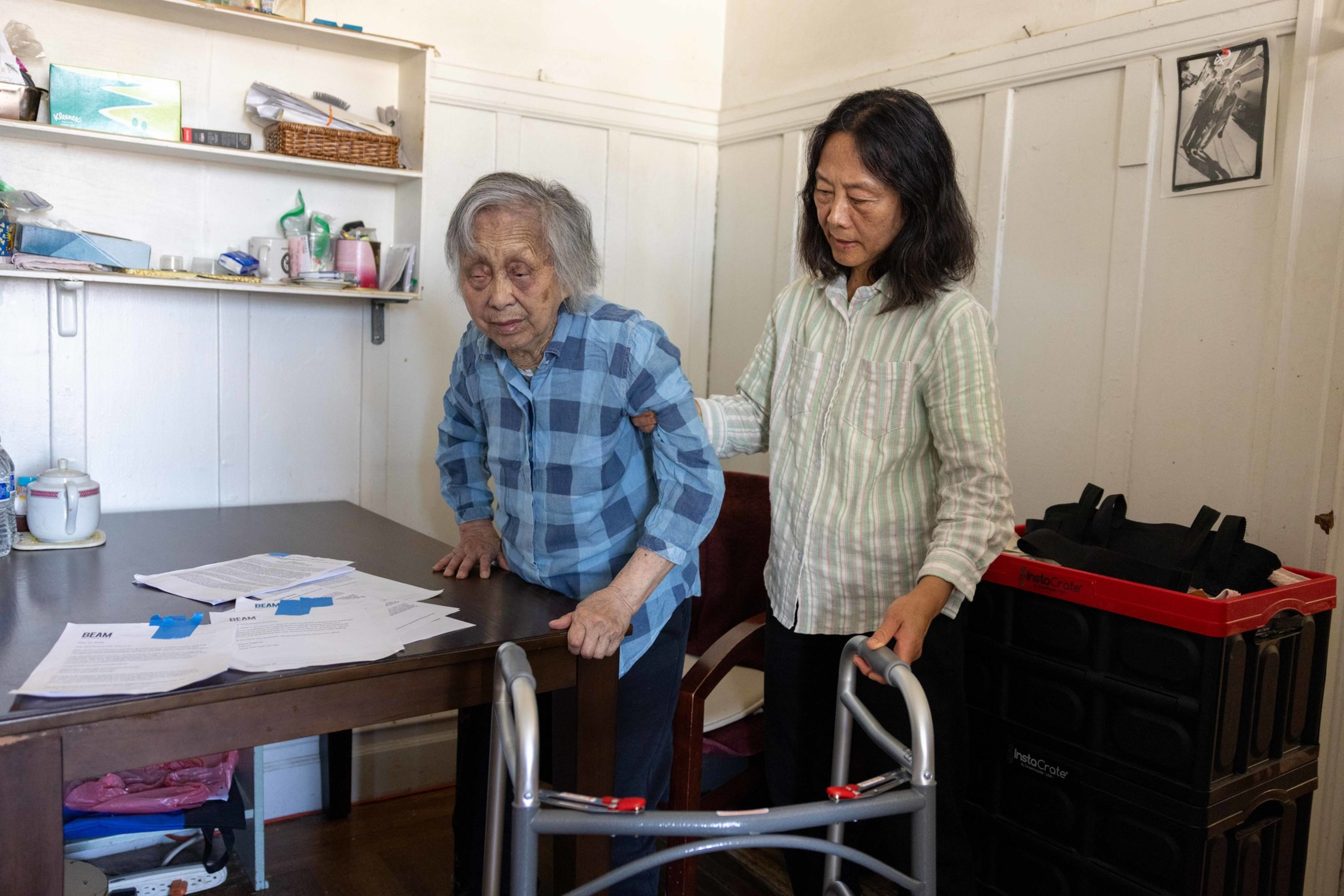 An elderly woman is being assisted by a middle-aged woman, using a walker near a table with scattered papers in a modest room.