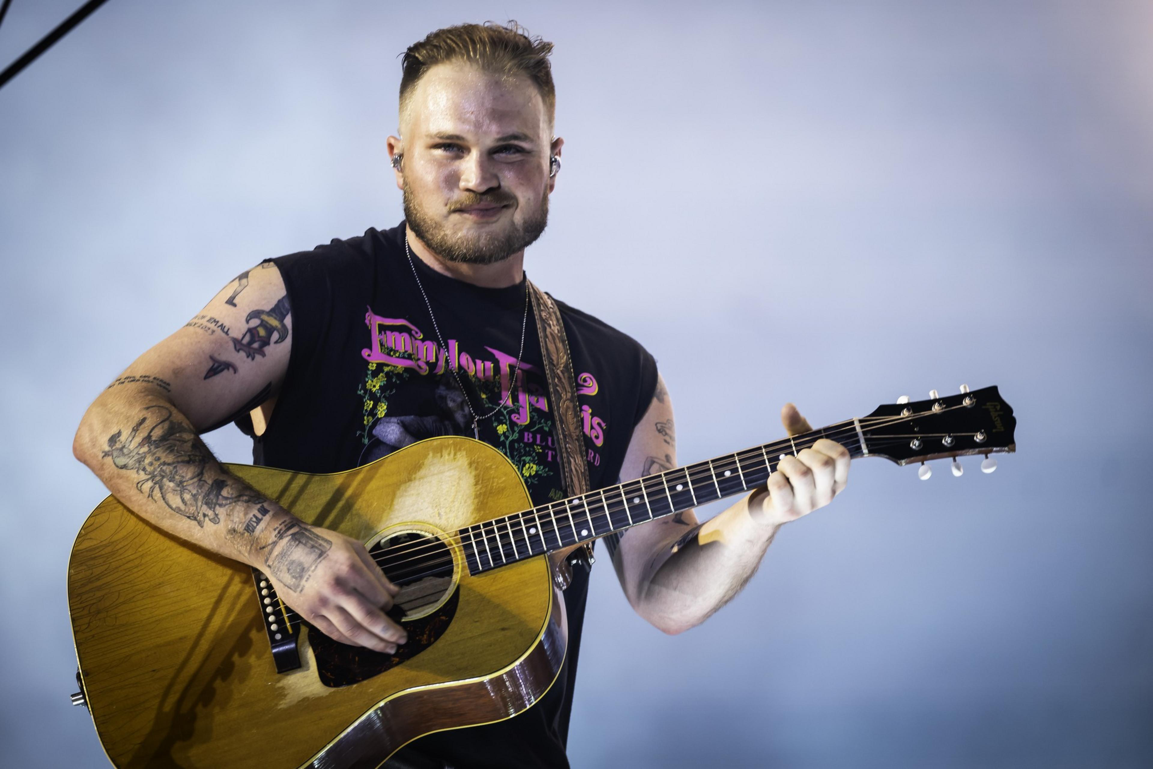 A man with tattoos plays an acoustic guitar. He wears a sleeveless black shirt with colorful text, and the background is softly lit, giving a serene atmosphere.