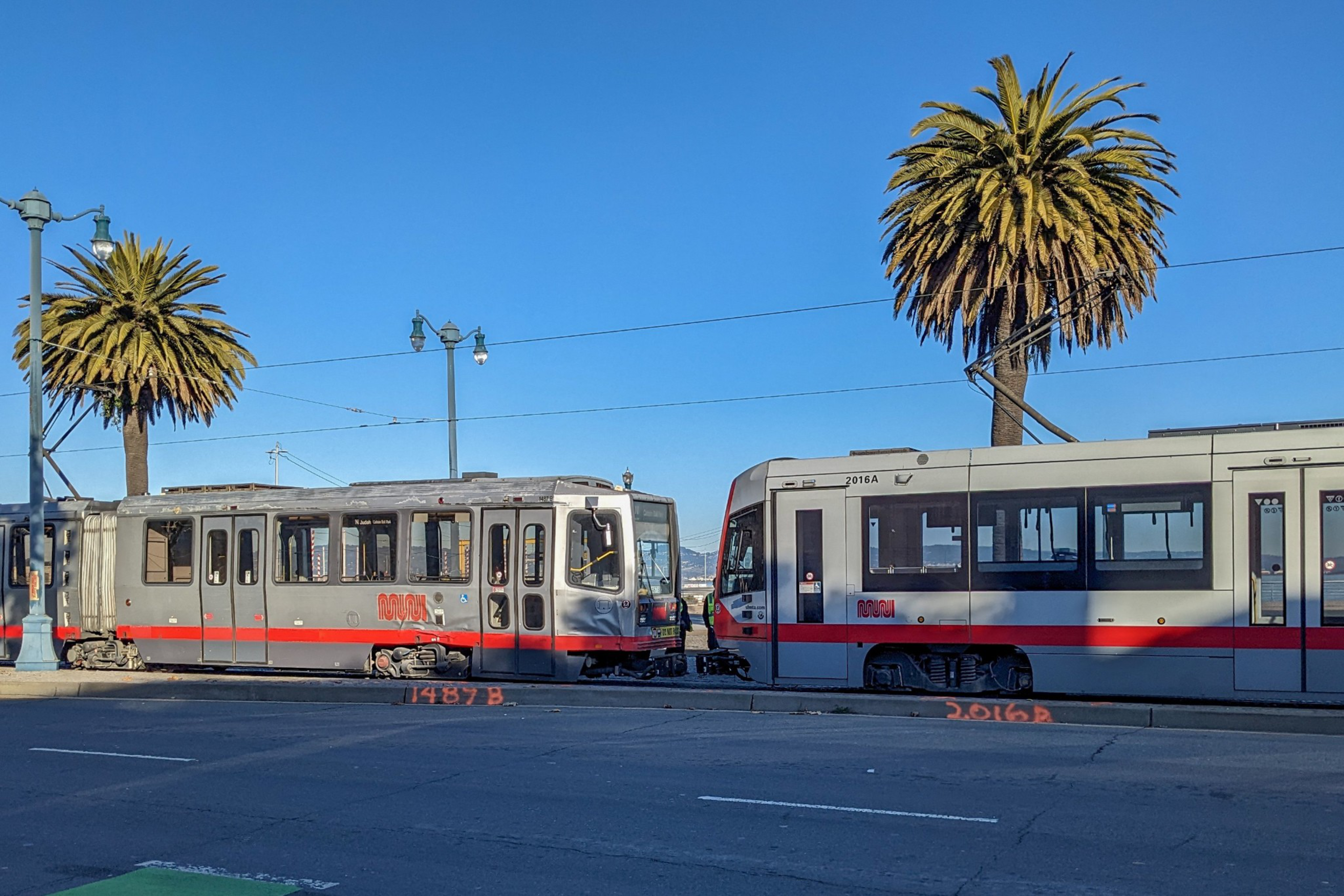 Two silver and red light rail trains face each other on tracks by a street, with two tall palm trees and a clear blue sky in the background.