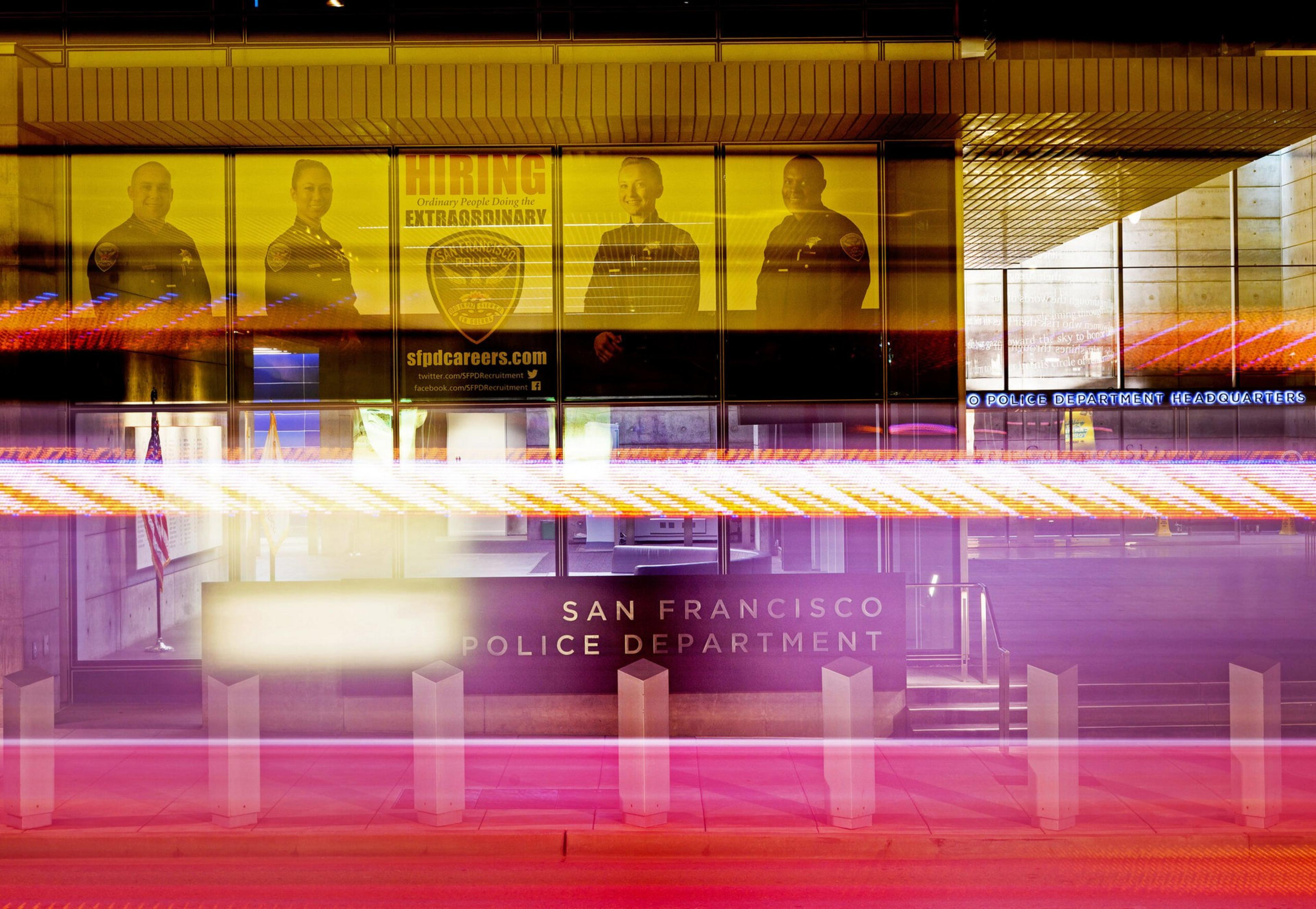 San Francisco Police Department building with a hiring poster displaying officers, streaks of colorful light in front, and the department's sign visible.