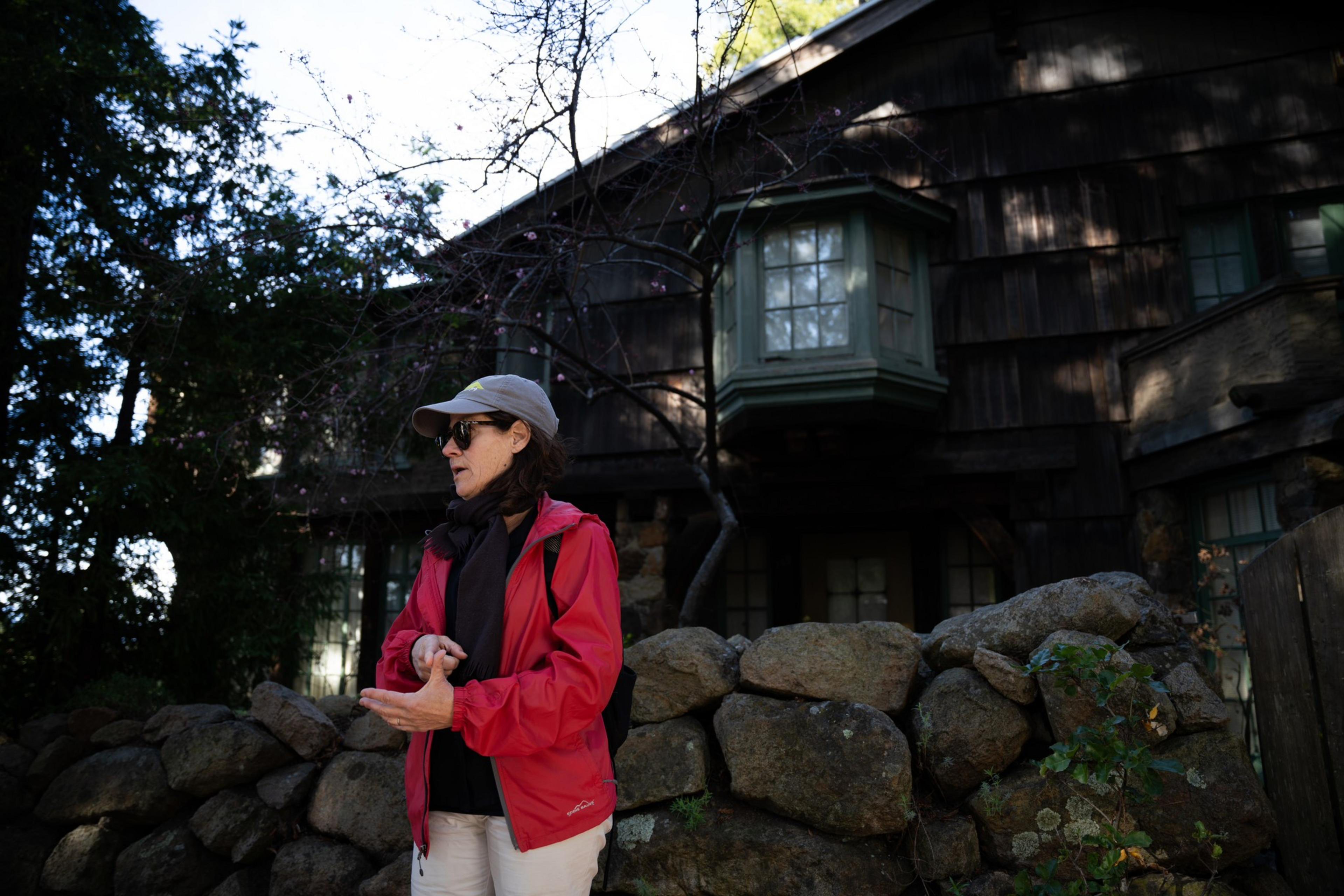 A person in a red jacket and cap stands near a stone wall, gesturing with their hands. Behind them is a rustic building with a large bay window.