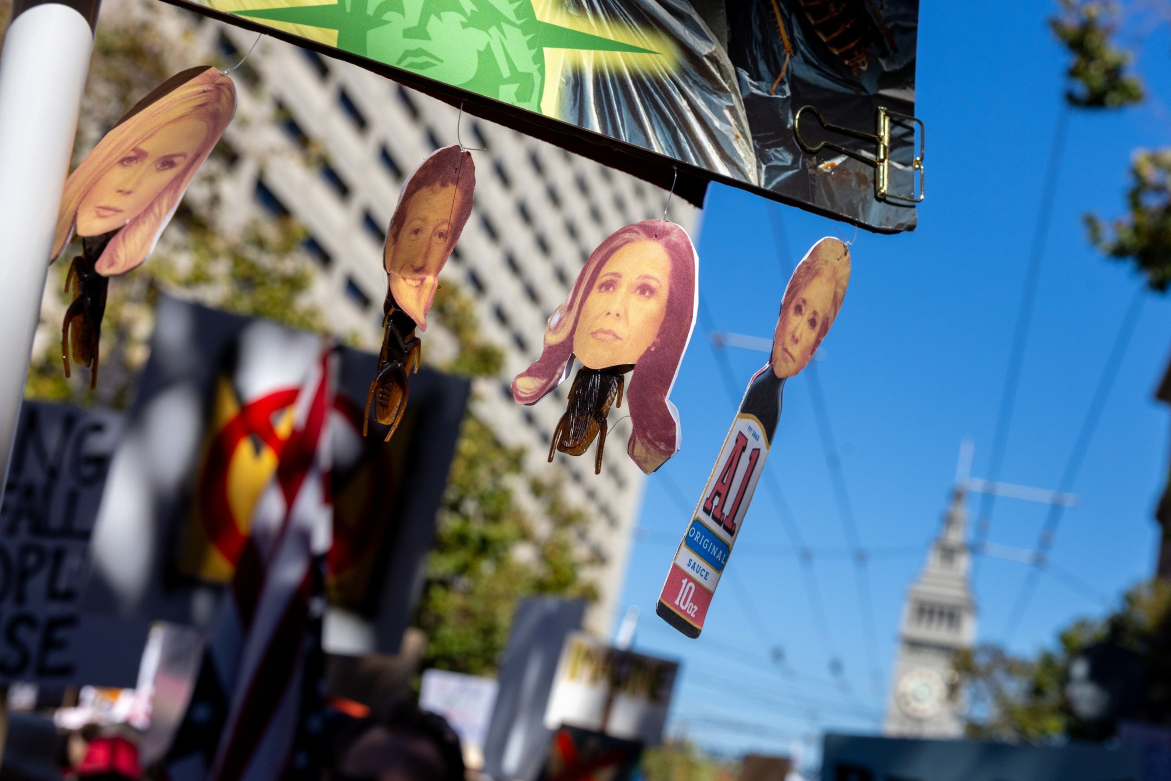 Cutout faces of various people hang from strings attached to an umbrella, with a crowd and a clock tower visible in the background on a bright day.