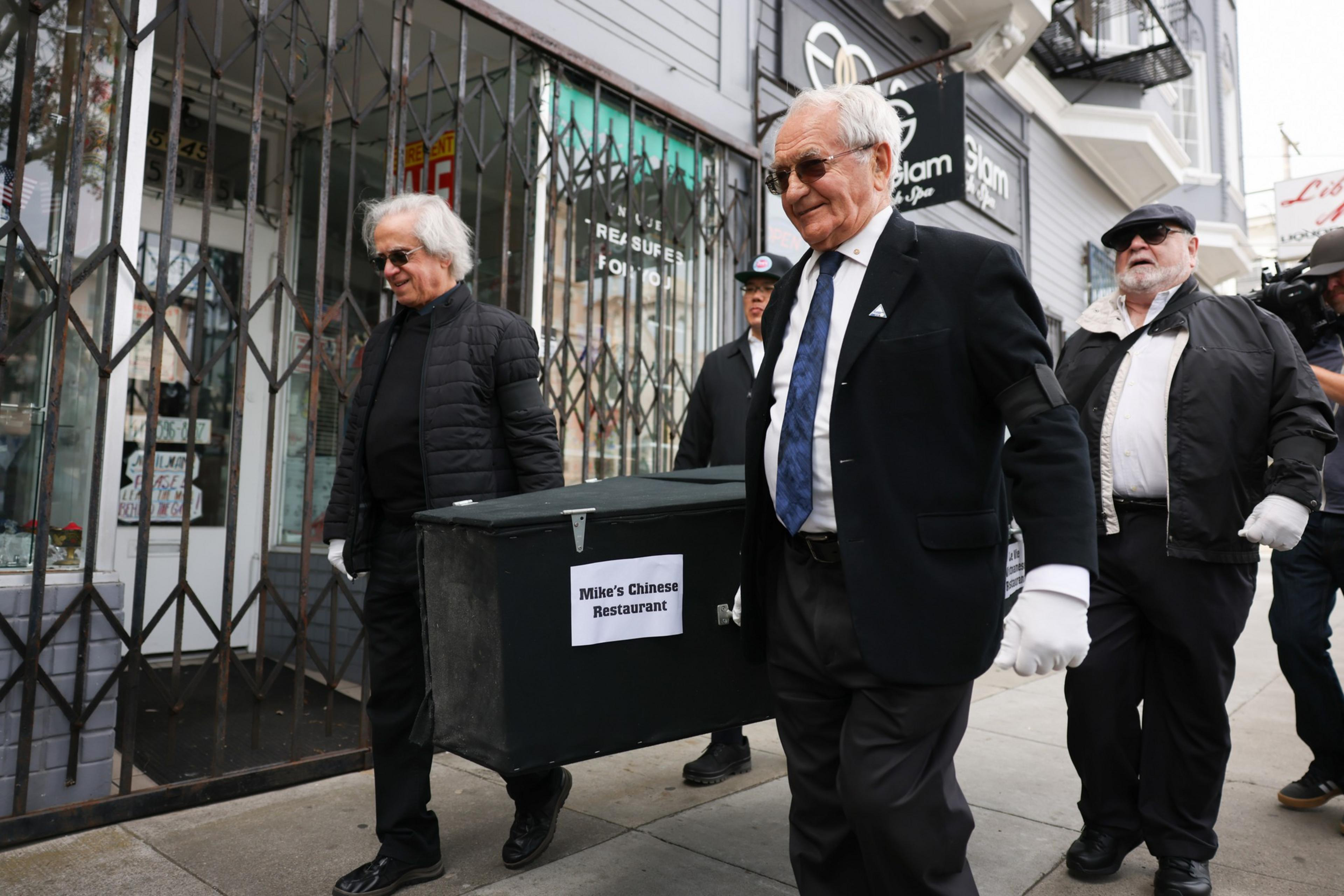 Four elderly men, dressed in black suits and gloves, are carrying a coffin labeled "Mike's Chinese Restaurant" down a city sidewalk.