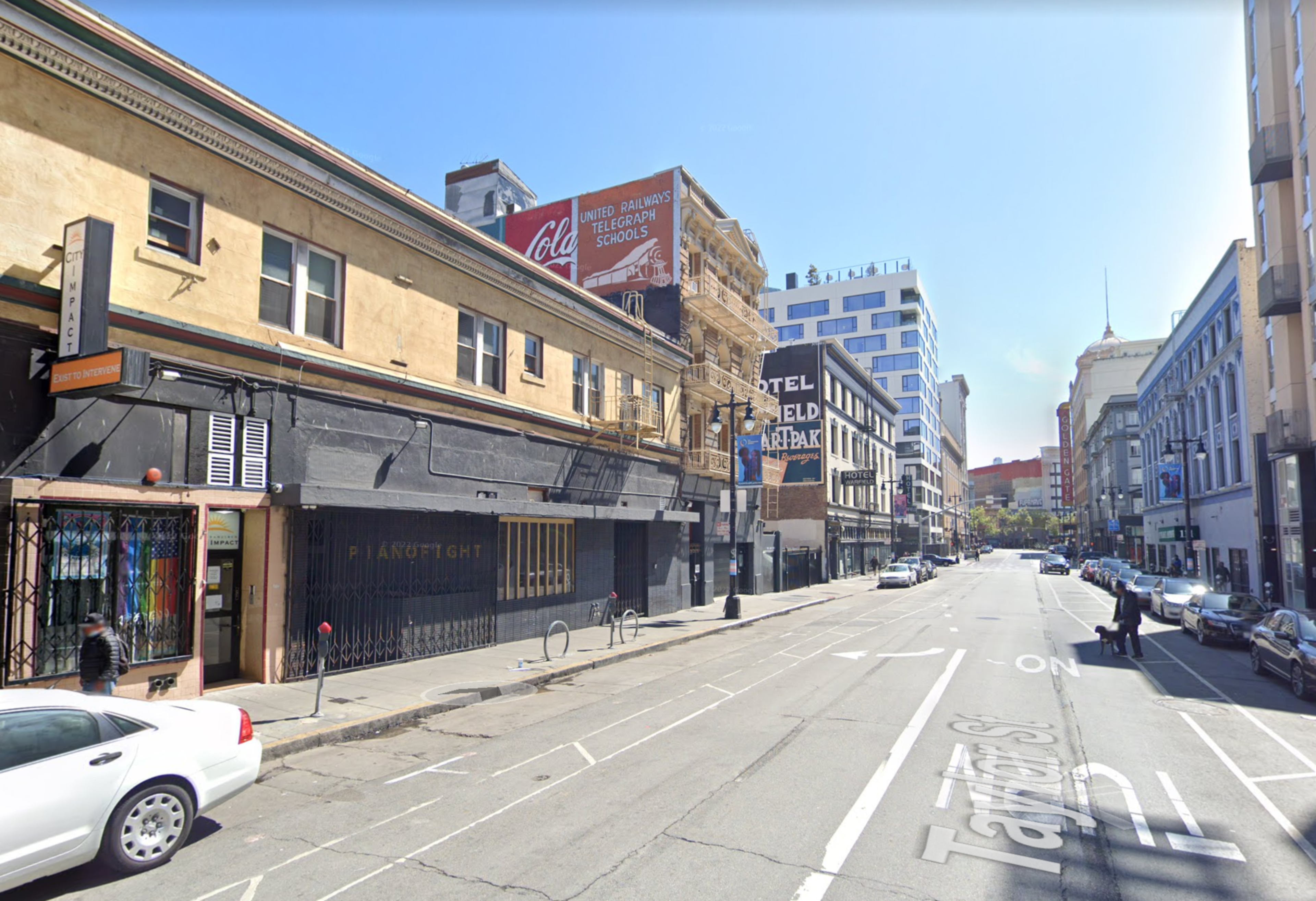 A quiet city street lined with parked cars, older buildings with faded signs, and a few pedestrians walking on the sidewalks under a clear blue sky.