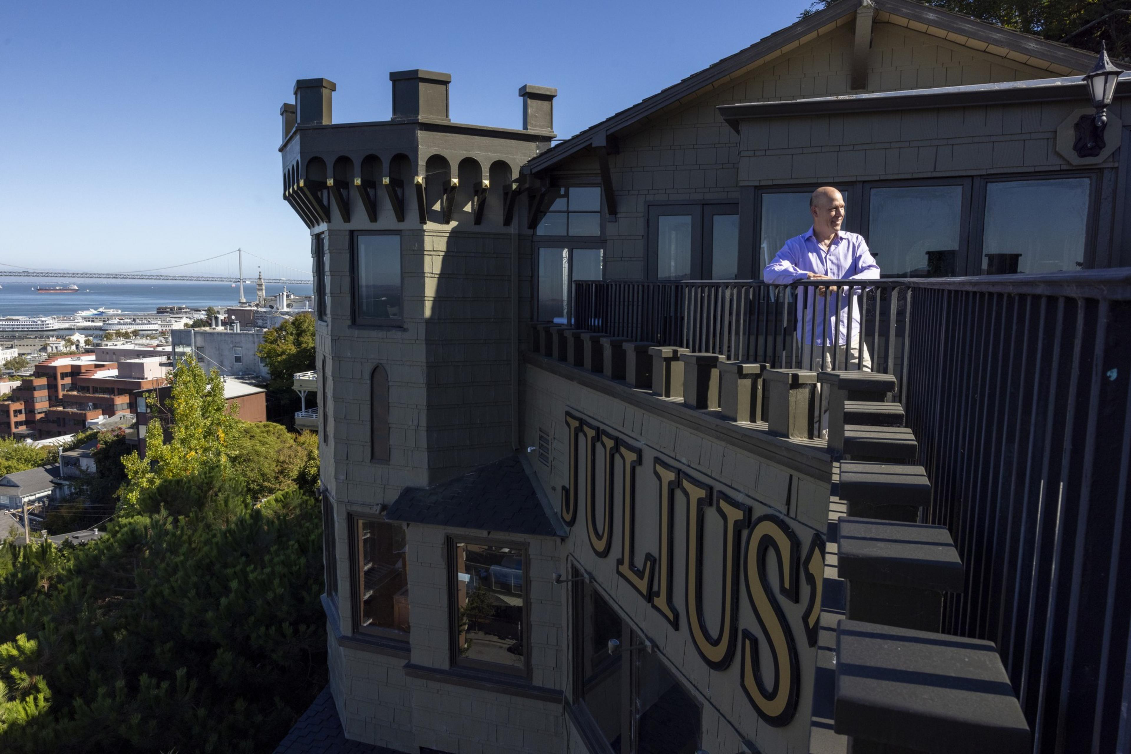 A man stands on a balcony with "Julius'" inscribed below him, overlooking a cityscape with a distant bridge and water visible under a clear blue sky.