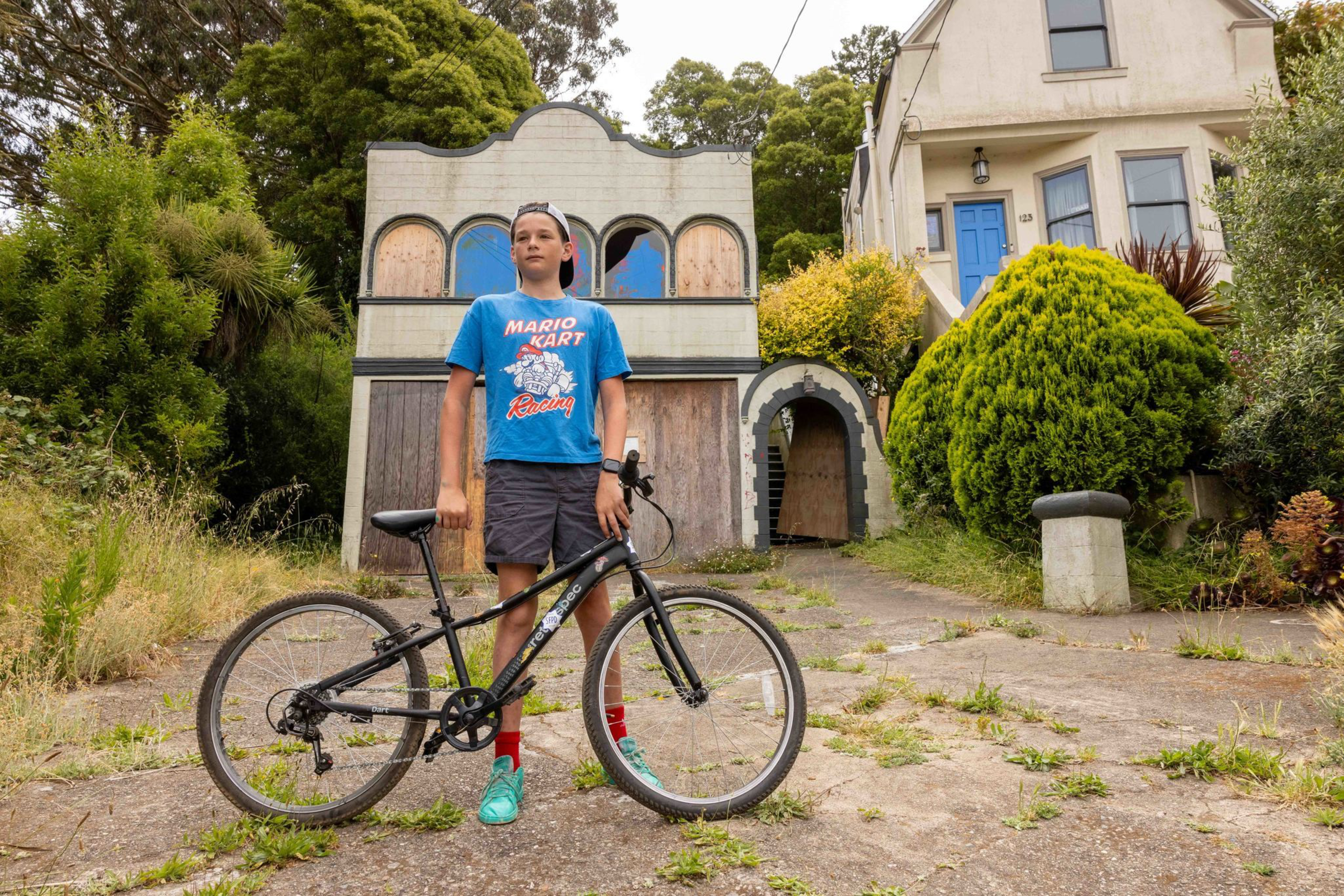 A boy in a blue Mario Kart shirt stands with a black bicycle on a cracked driveway in front of an old, boarded-up building and a house with a blue door.
