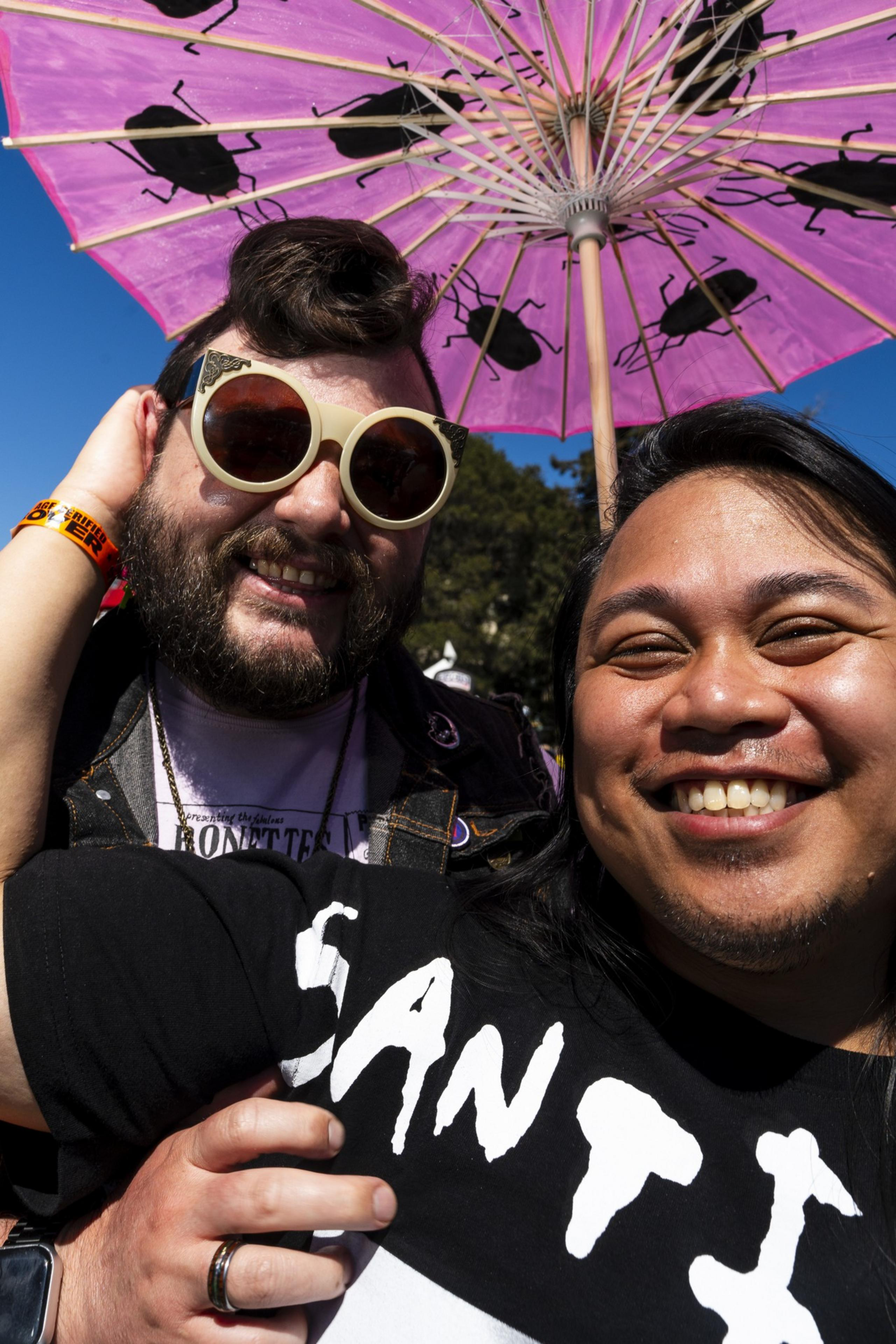 Two people pose happily under a pink parasol with black beetle designs. One wears large sunglasses and a denim vest, while the other has long hair and a T-shirt.