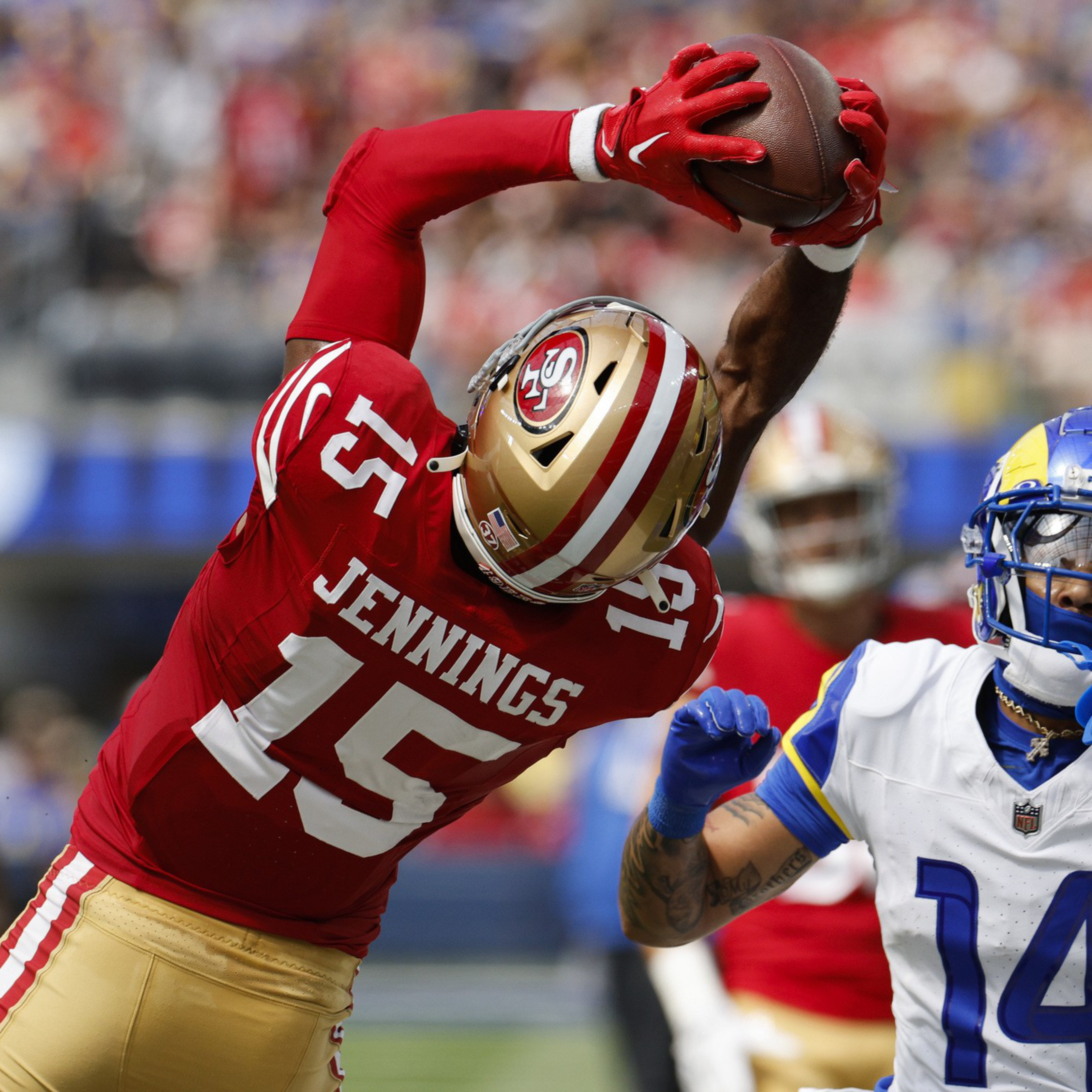 A football player in a red and gold uniform labeled Jennings 15 leaps to catch the ball while a player in a white and blue uniform attempts to defend.