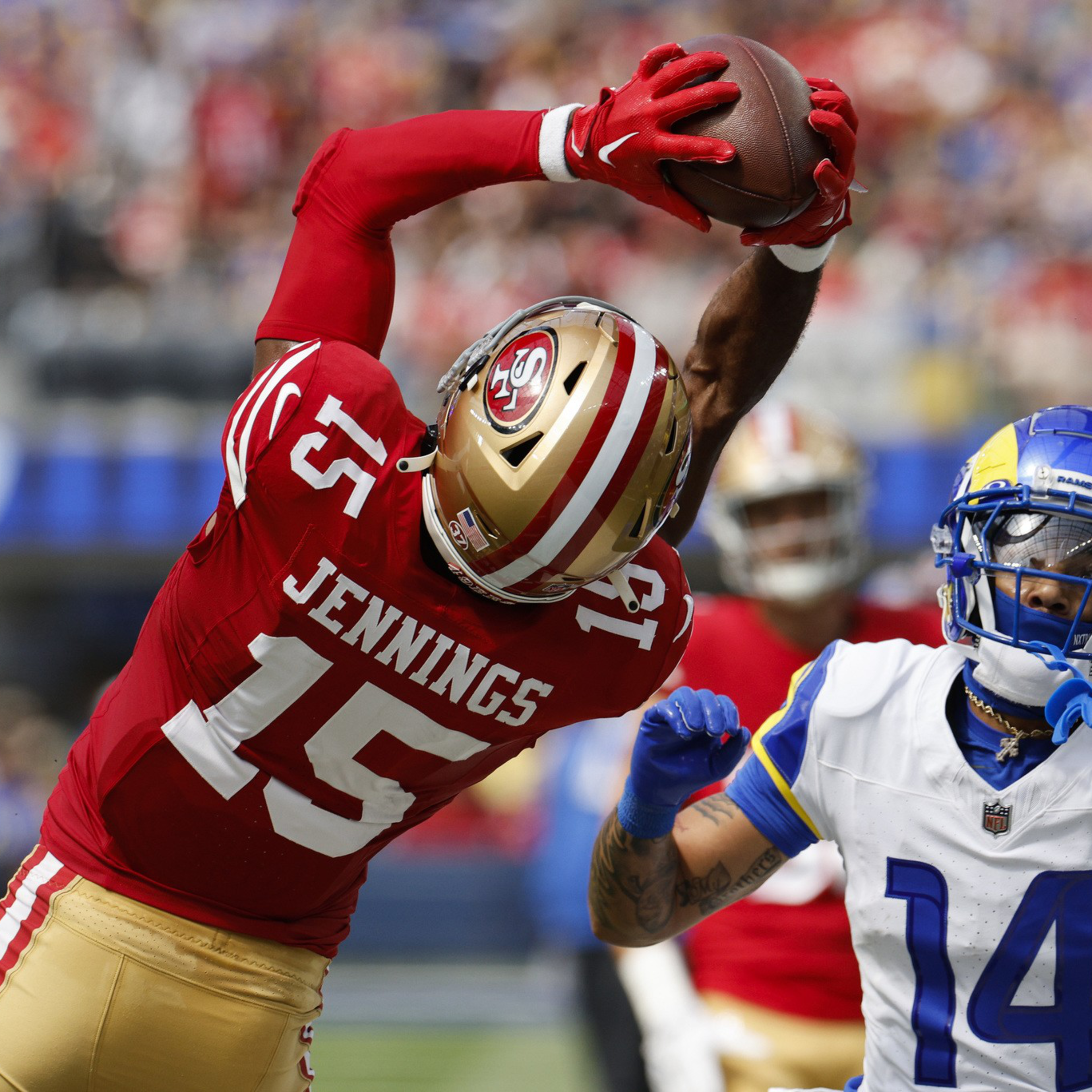 A football player in a red and gold uniform labeled Jennings 15 leaps to catch the ball while a player in a white and blue uniform attempts to defend.