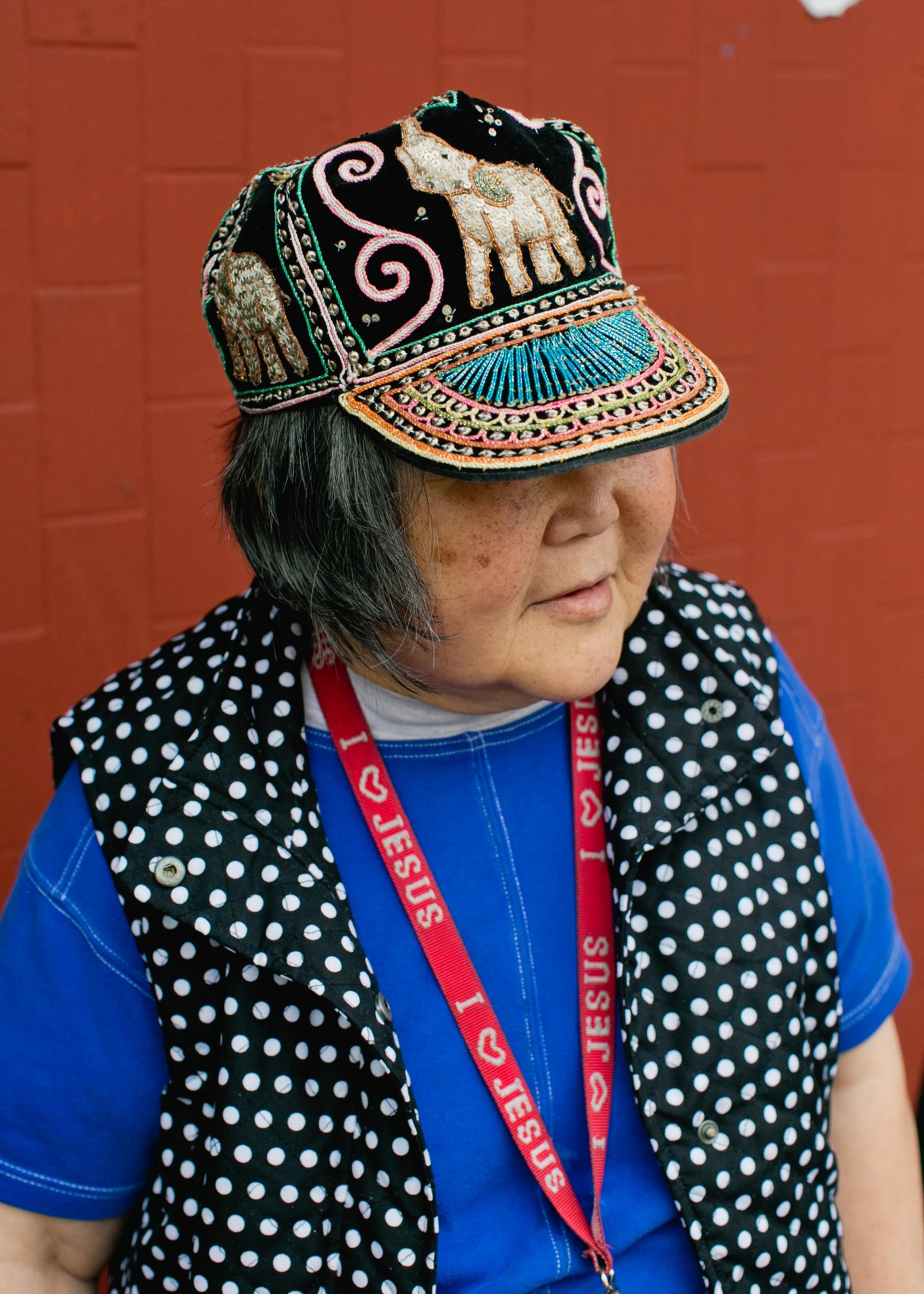 A person wears a colorful embroidered hat with elephants, a blue shirt, a black and white polka-dot vest, and a red lanyard reading “I ♥ JESUS.”