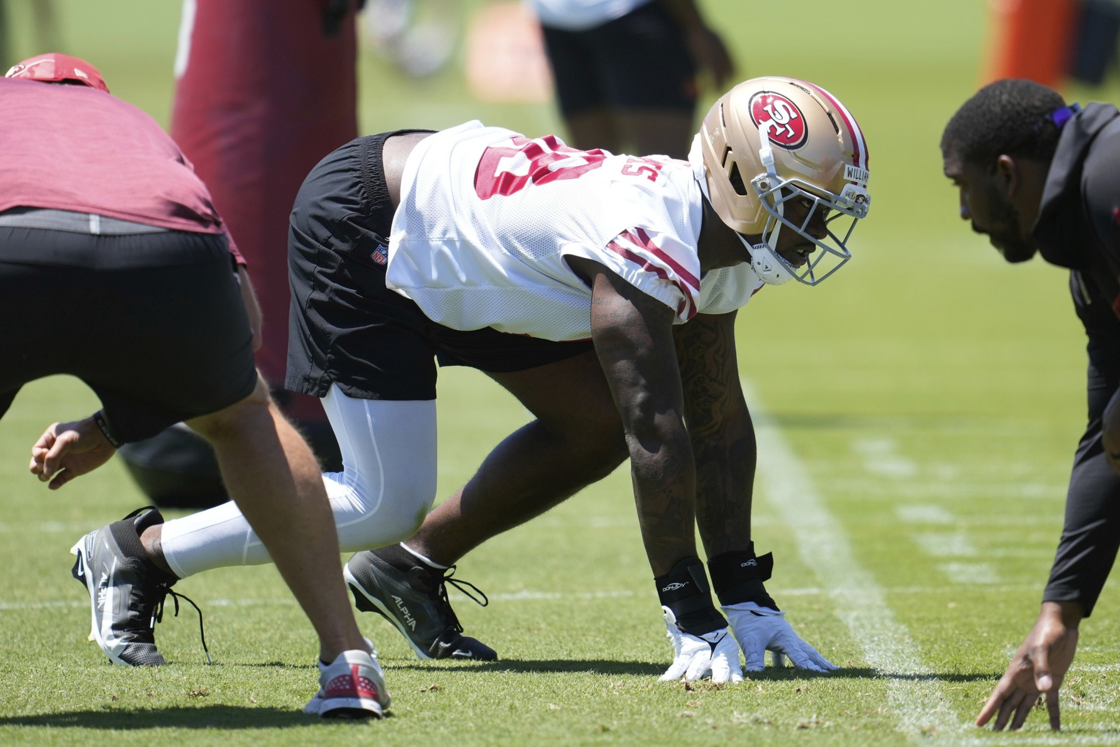 Two football players face each other crouched in a stance on a practice field, wearing helmets and team gear under a sunny sky.