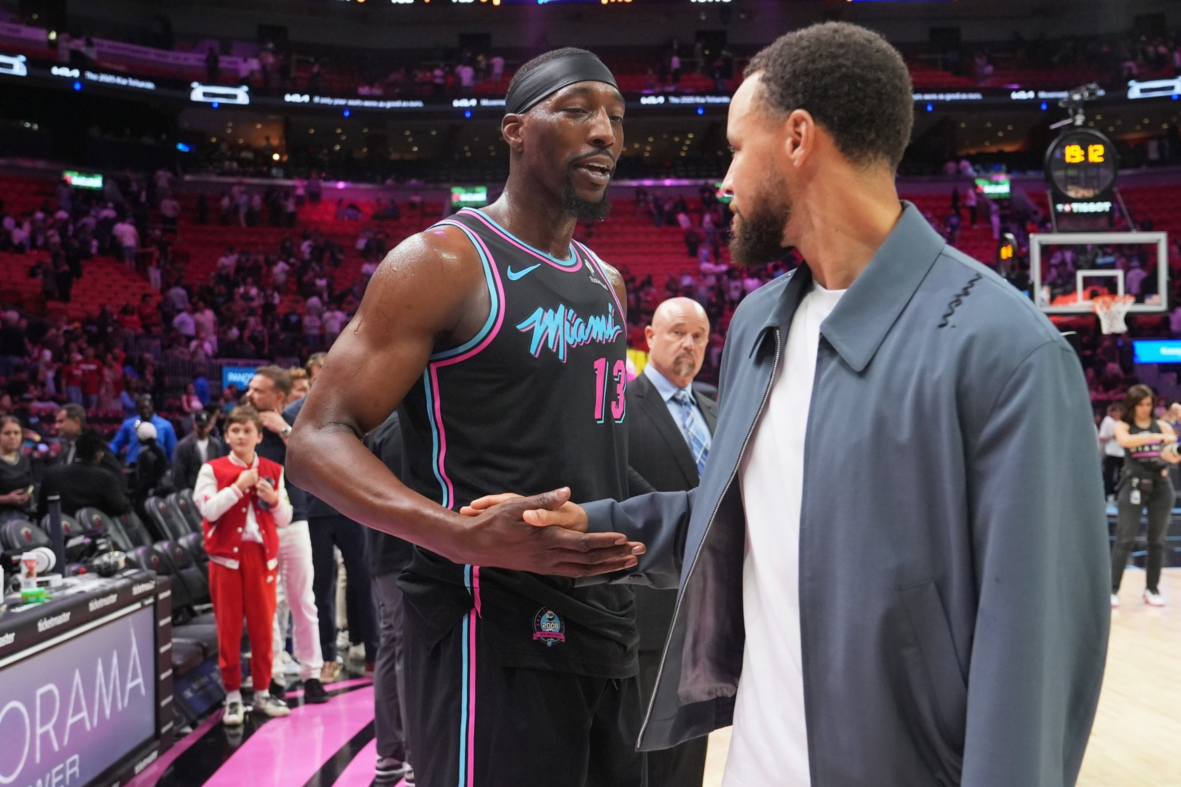 A basketball player in a Miami Heat uniform shakes hands with a man in a blue jacket on a basketball court with spectators in the background.